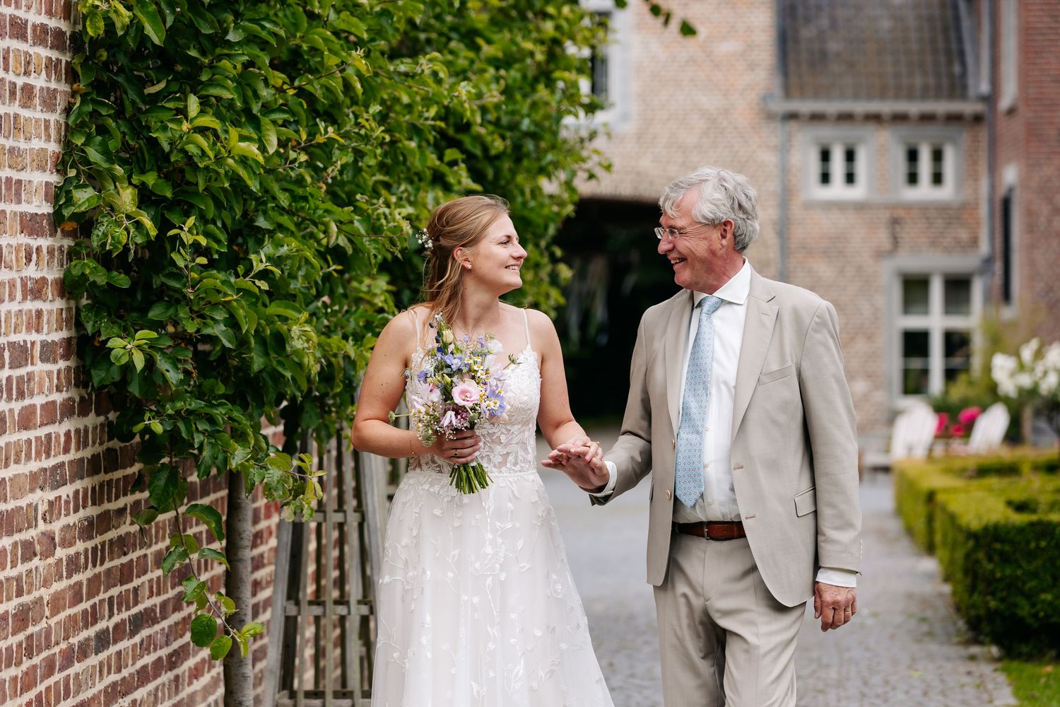 Bride holding a bouquet walking hand in hand with her father along an ivy-covered brick wall.