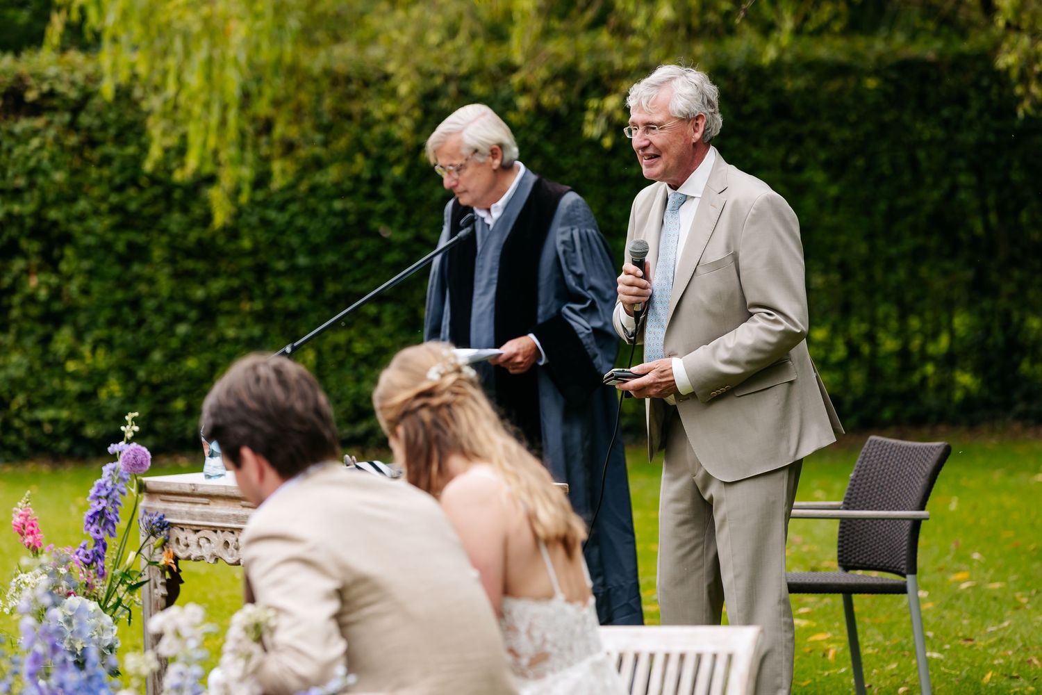 Officiant and guest laughing during an outdoor wedding ceremony in a lush green garden with floral decorations.