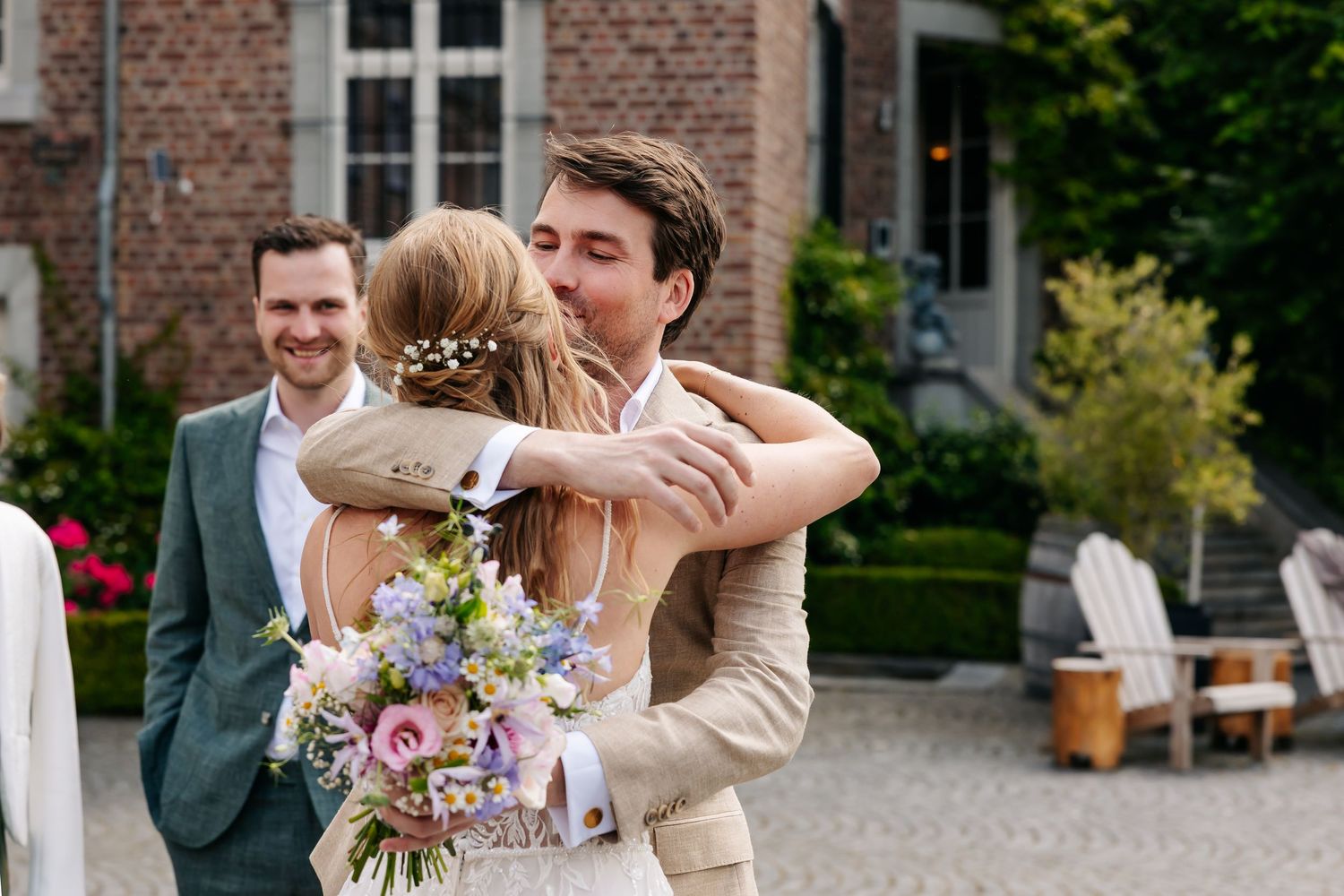 Bride in lace gown with floral bouquet embracing guest at outdoor wedding ceremony.