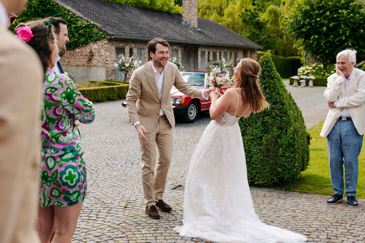 Groom in tan suit greeting bride in white wedding dress outside brick manor with red vintage car.