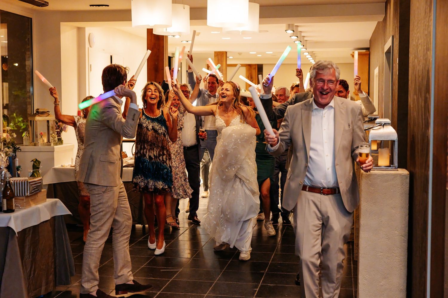 Bride and groom entering reception hall with glowing light sticks amid cheering guests.