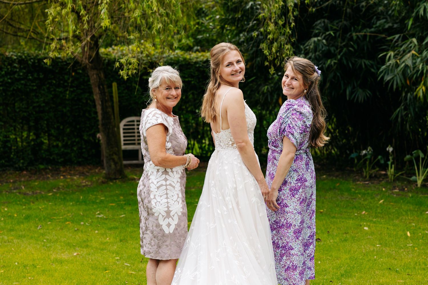 Bride in lace wedding gown posing with two smiling women in garden setting.