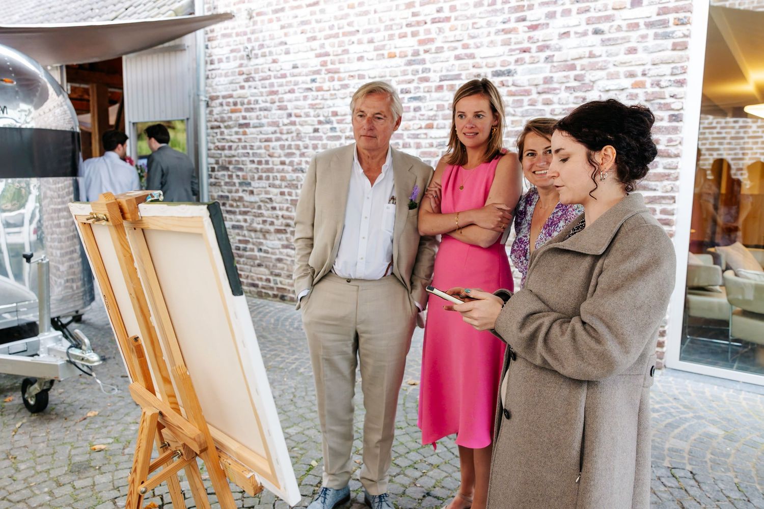 Group of elegantly dressed guests examining a painting on an easel at an outdoor wedding venue.