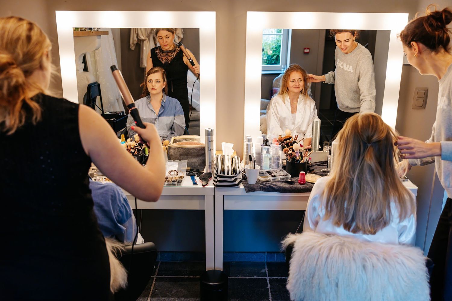 Bridal party getting hair and makeup done at a lit vanity mirror in a professional dressing room.