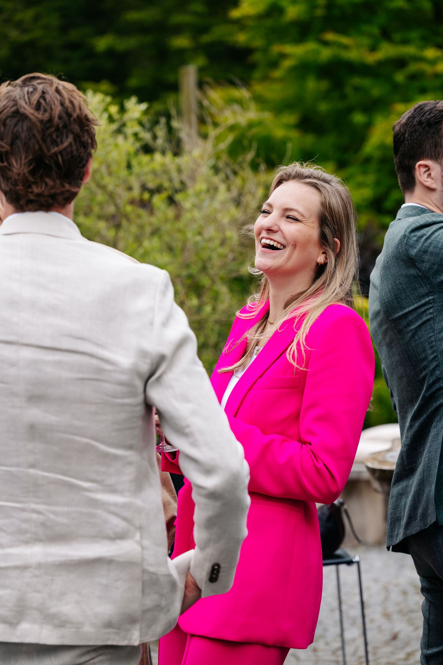 Woman in a bright pink suit laughing joyfully with guests at an outdoor wedding.
