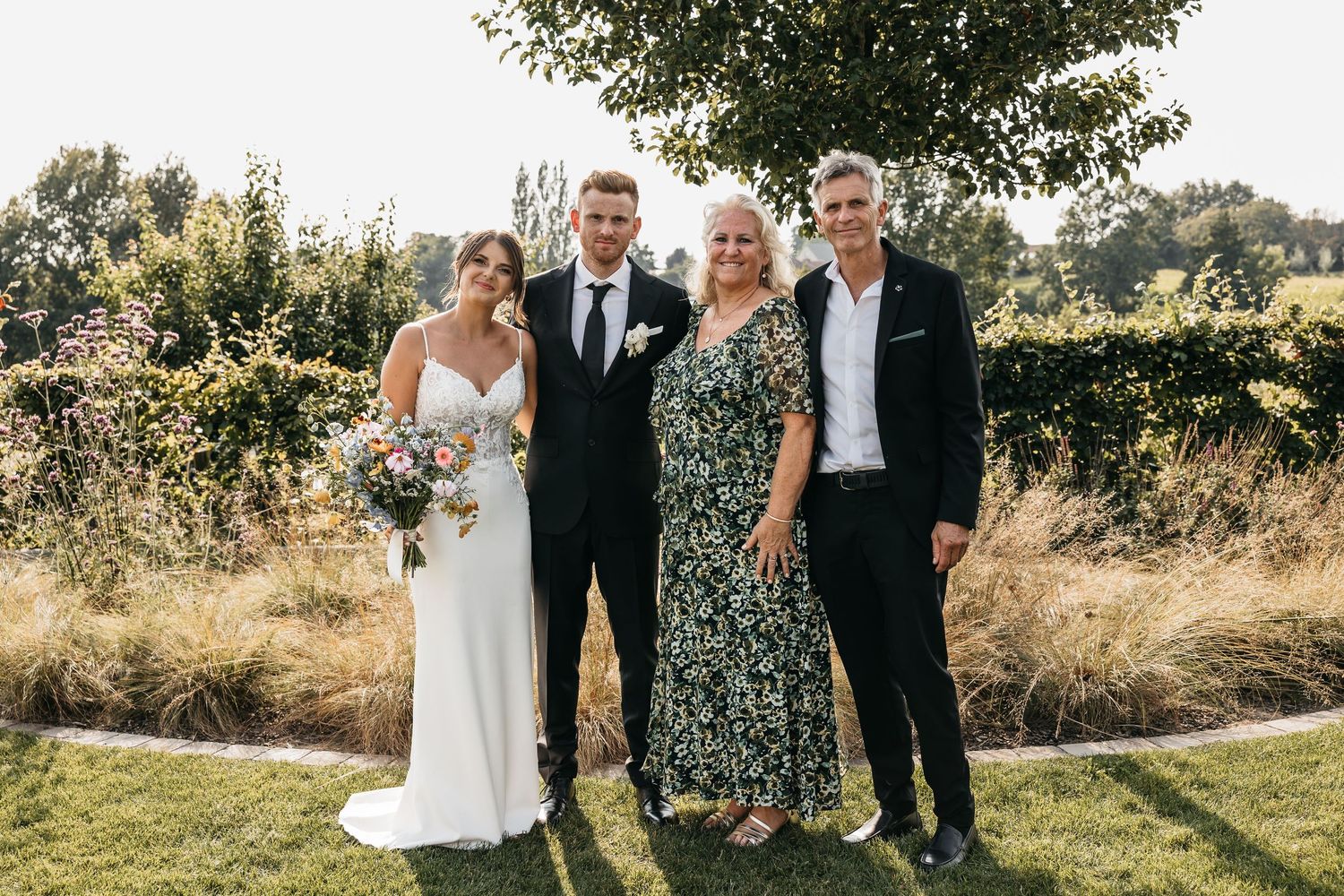 Bride and groom pose with two family members in a sunny garden setting, bride holding a colorful floral bouquet.