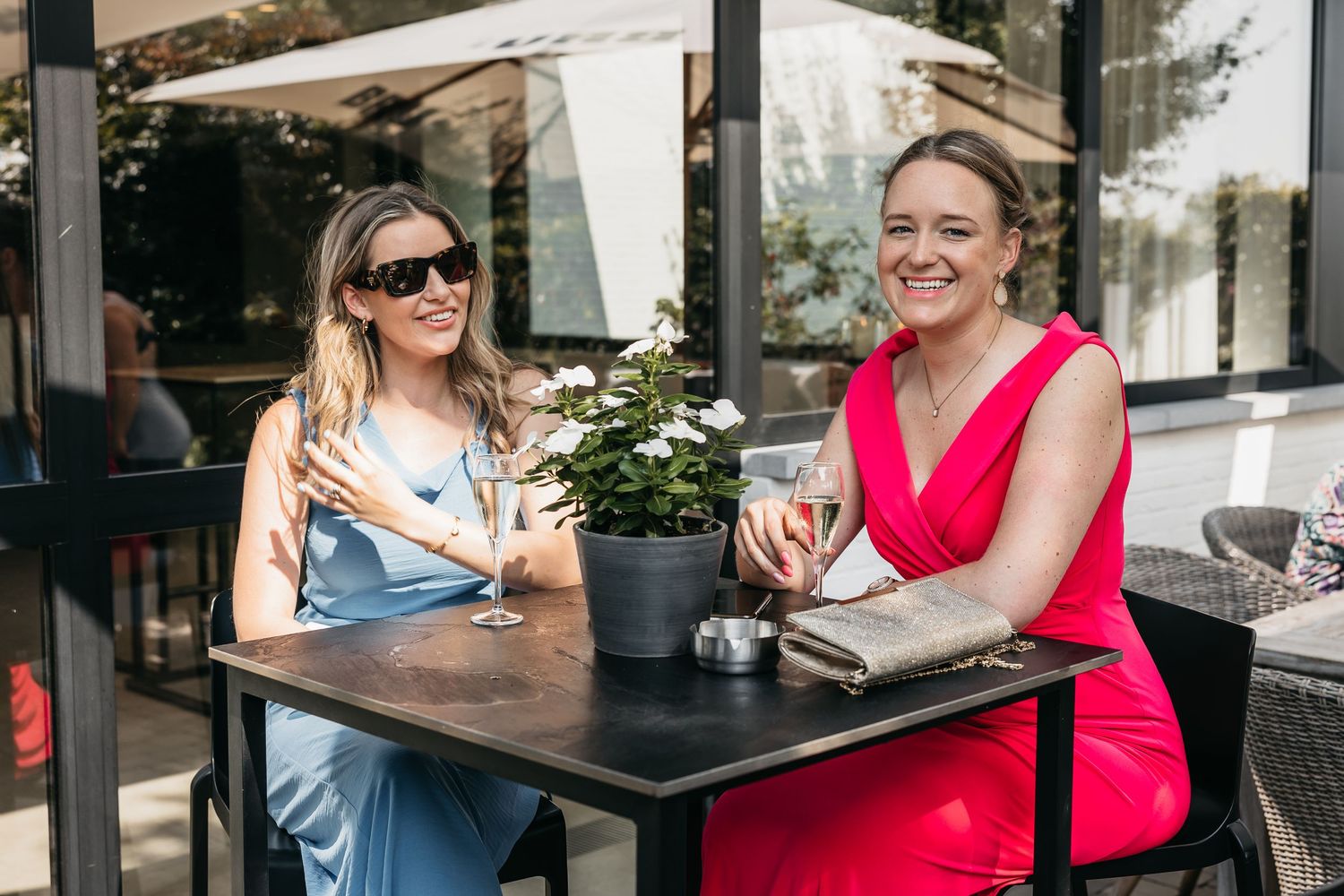 Two women in blue and pink dresses sitting at outdoor table with white flowers at wedding.