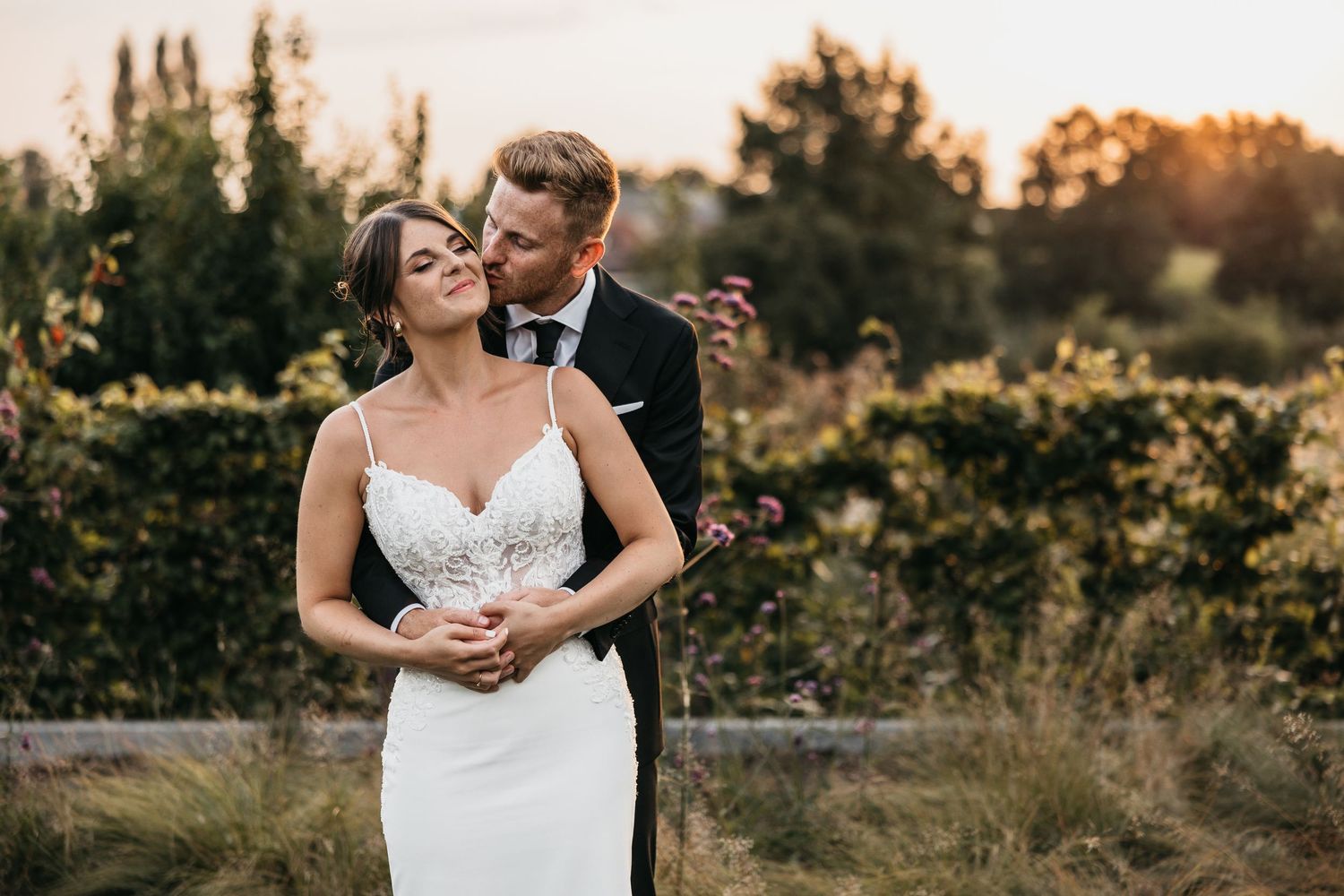 Groom in black suit kisses bride on the cheek while embracing her from behind in a golden sunset garden setting.