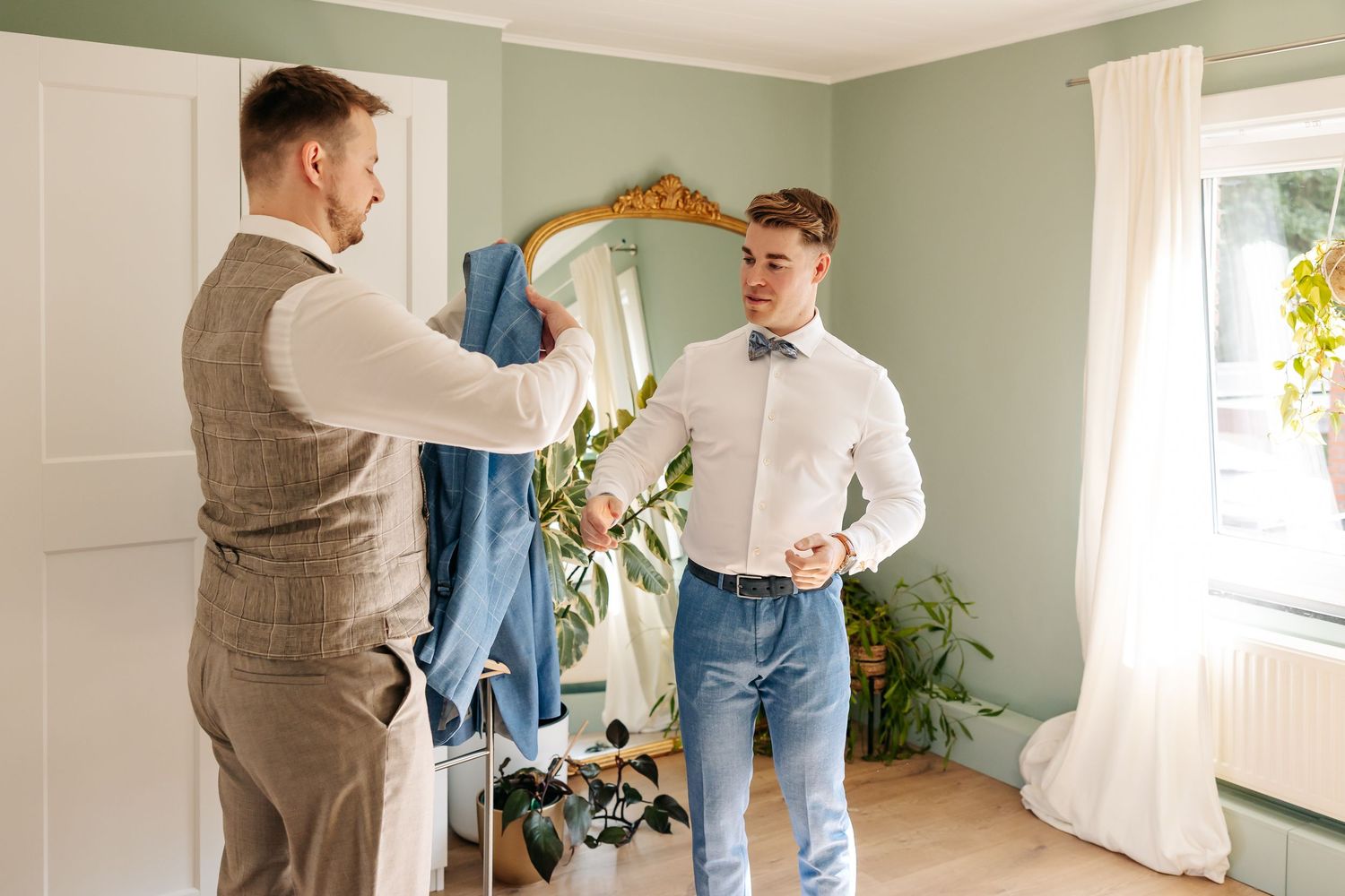 Groomsman presenting blue suit jacket to groom with bow tie in elegant room.