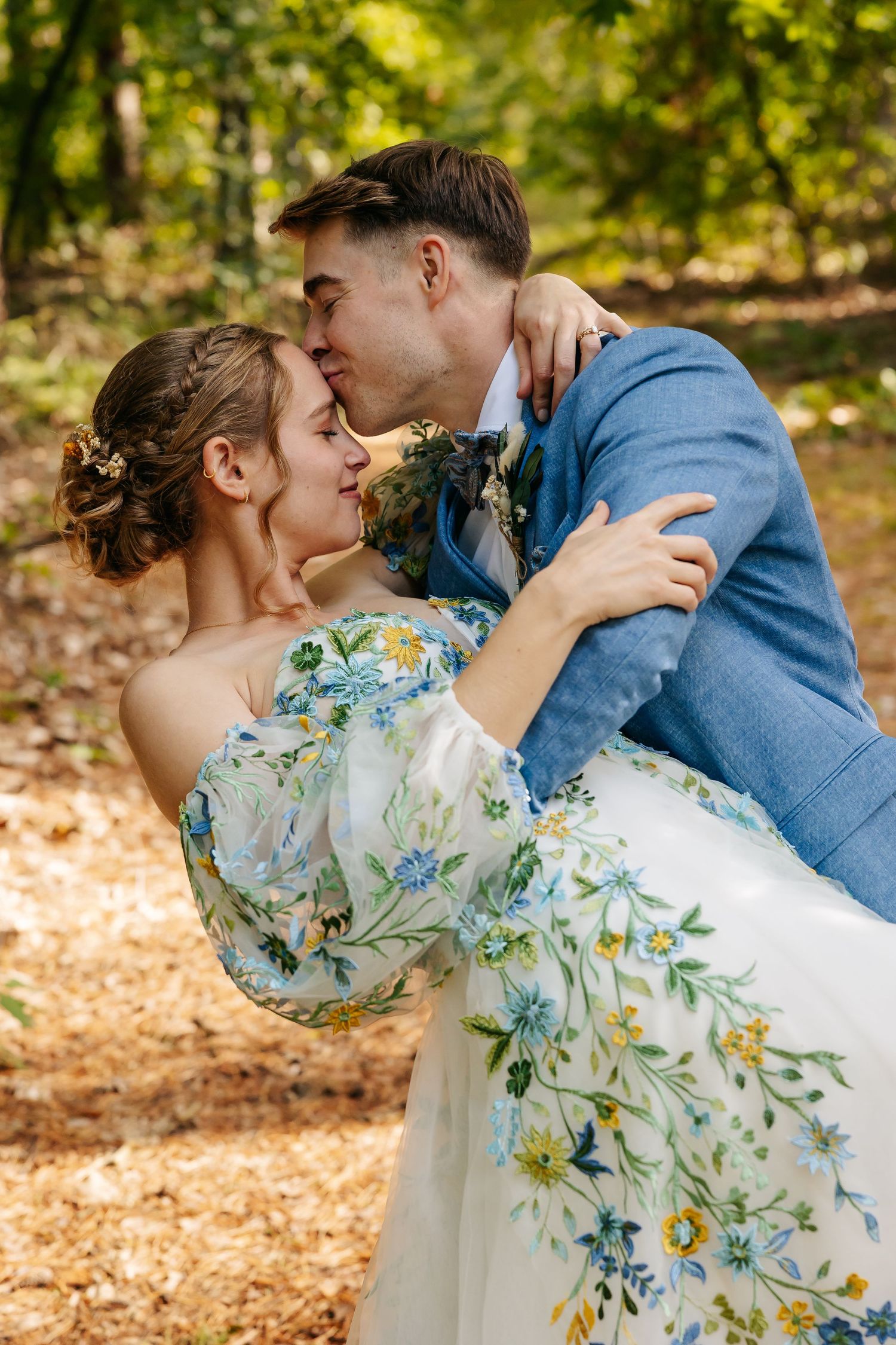 Groom kissing bride in floral wedding dress in sunlit forest.