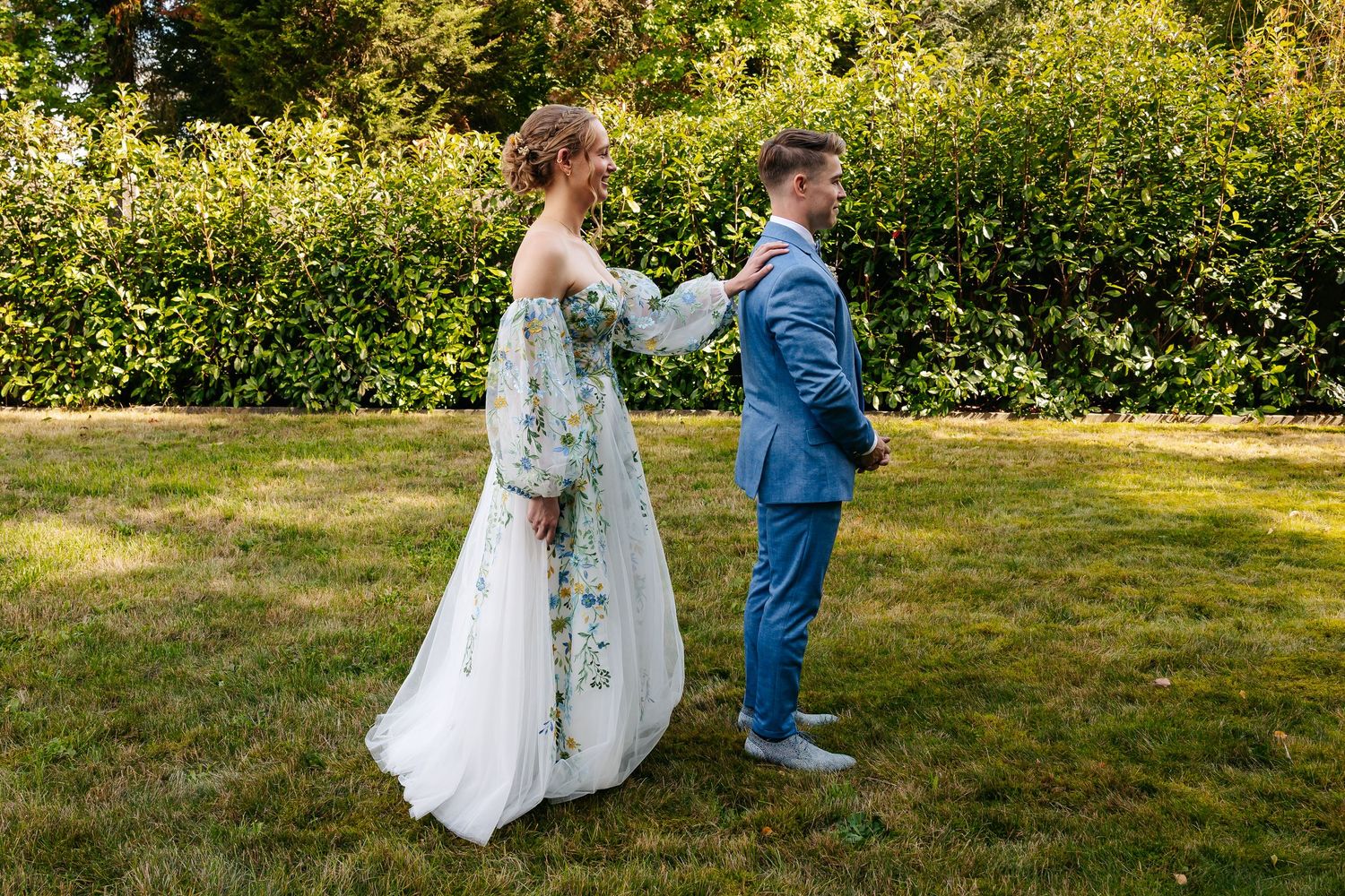 Bride in floral wedding dress adjusting grooms blue suit jacket in garden.