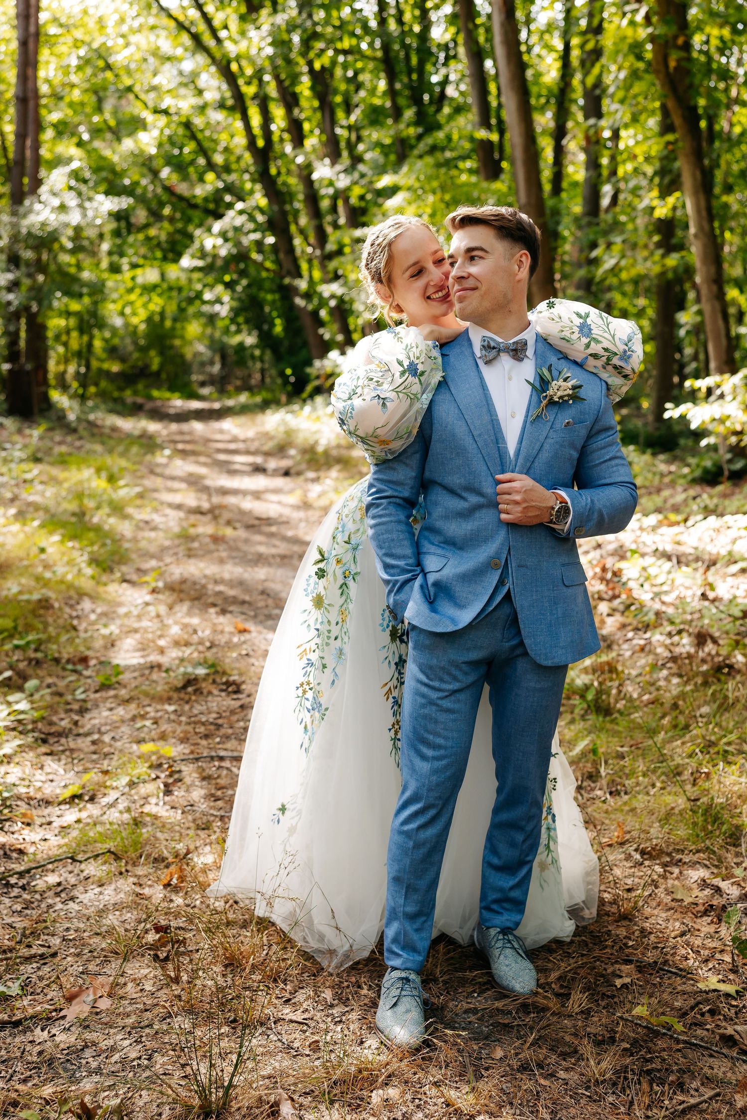 Bride in floral wedding dress embracing groom in blue suit on a sunlit forest trail.