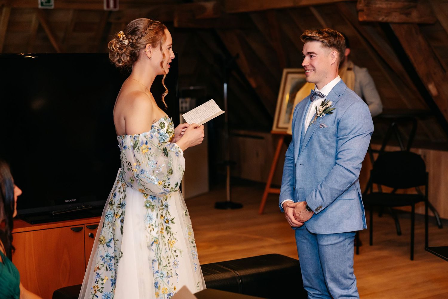 Bride reads wedding vows from paper to groom in blue suit during rustic barn ceremony.