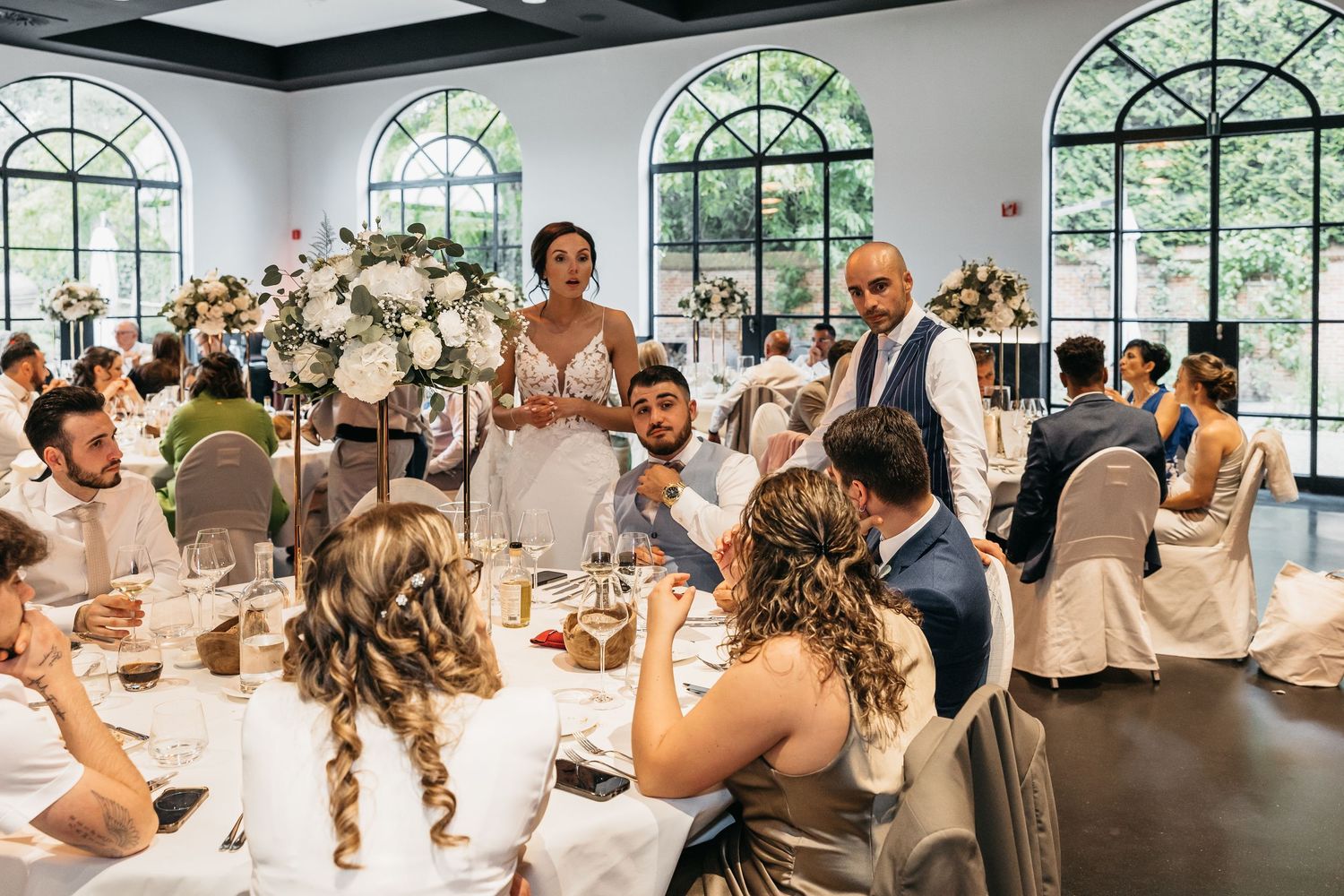 Bride and groom mingle with guests at elegant wedding reception featuring tall white floral centerpieces.