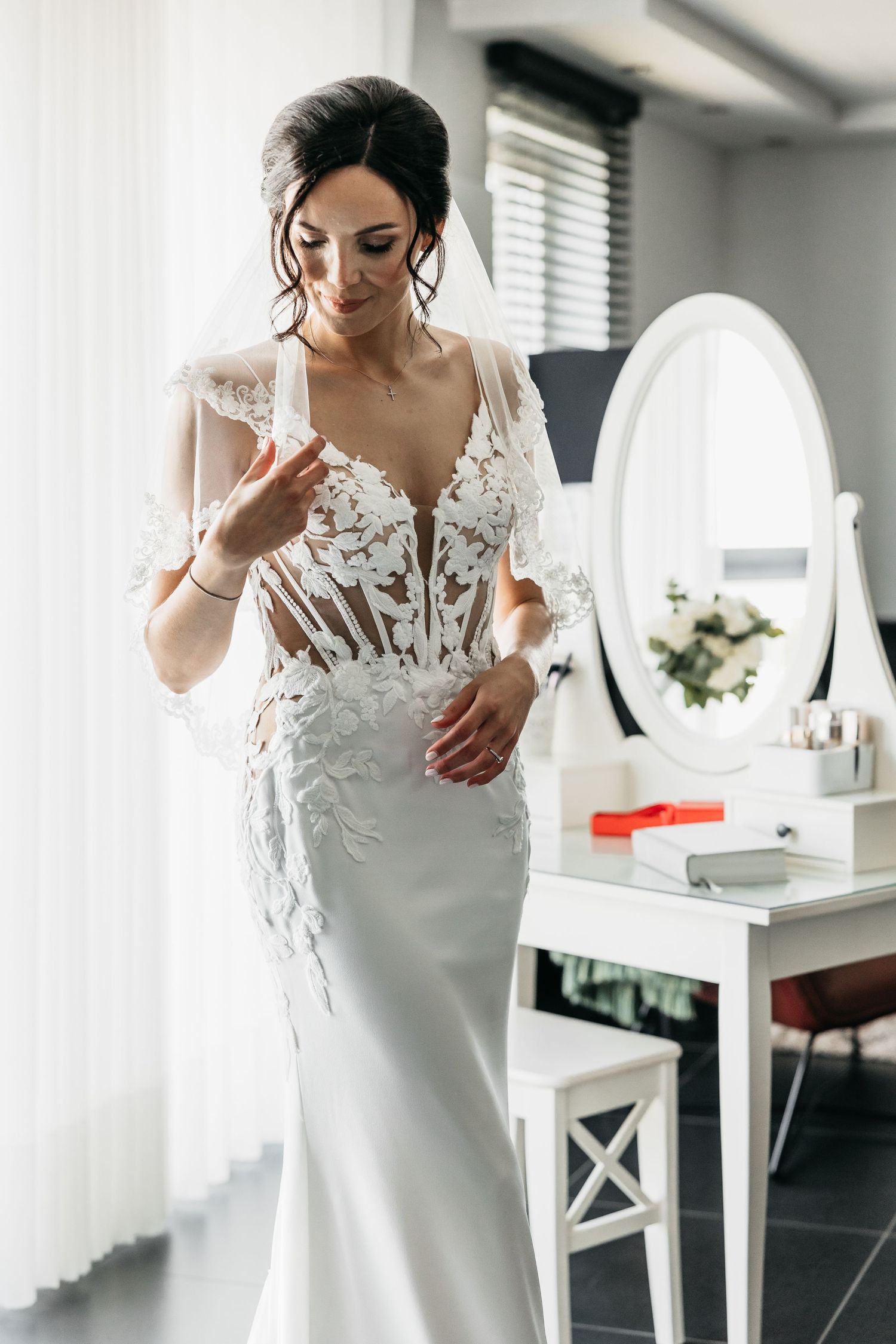 Stunning bride in elegant lace mermaid wedding gown standing near white oval vanity mirror indoors.