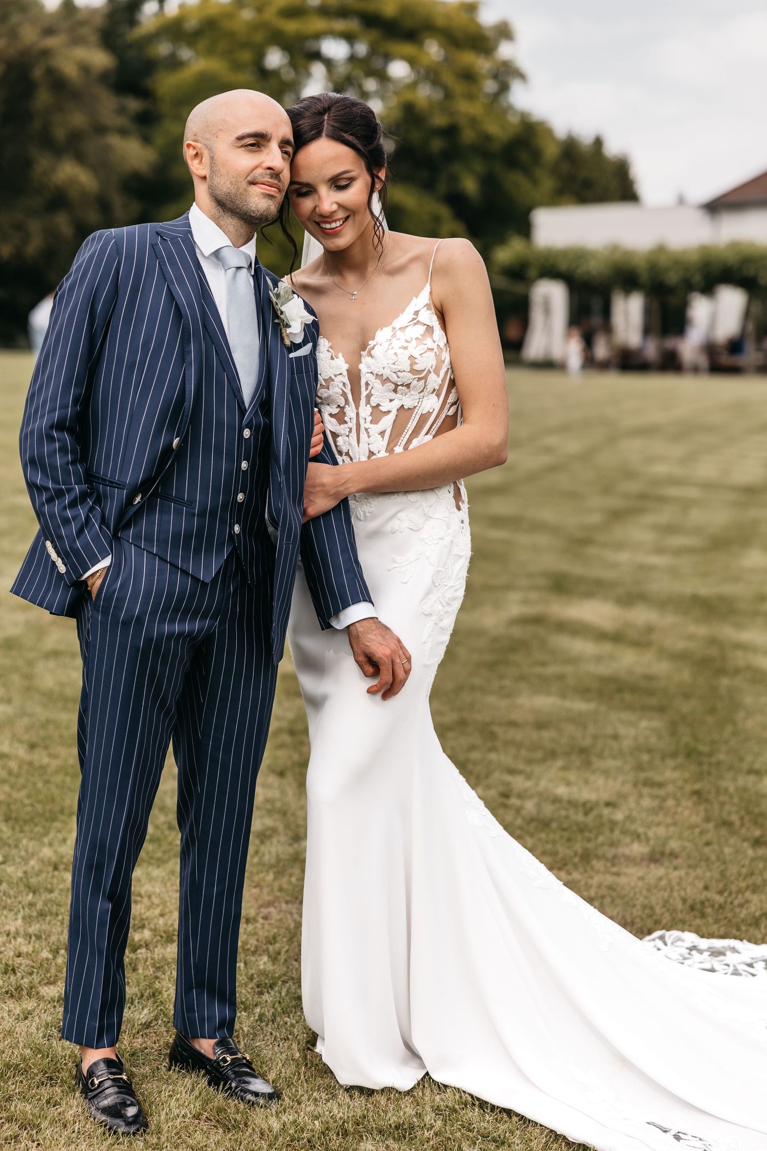 Bride in fitted lace gown embraces smiling bald groom in navy pinstripe suit on a garden lawn.