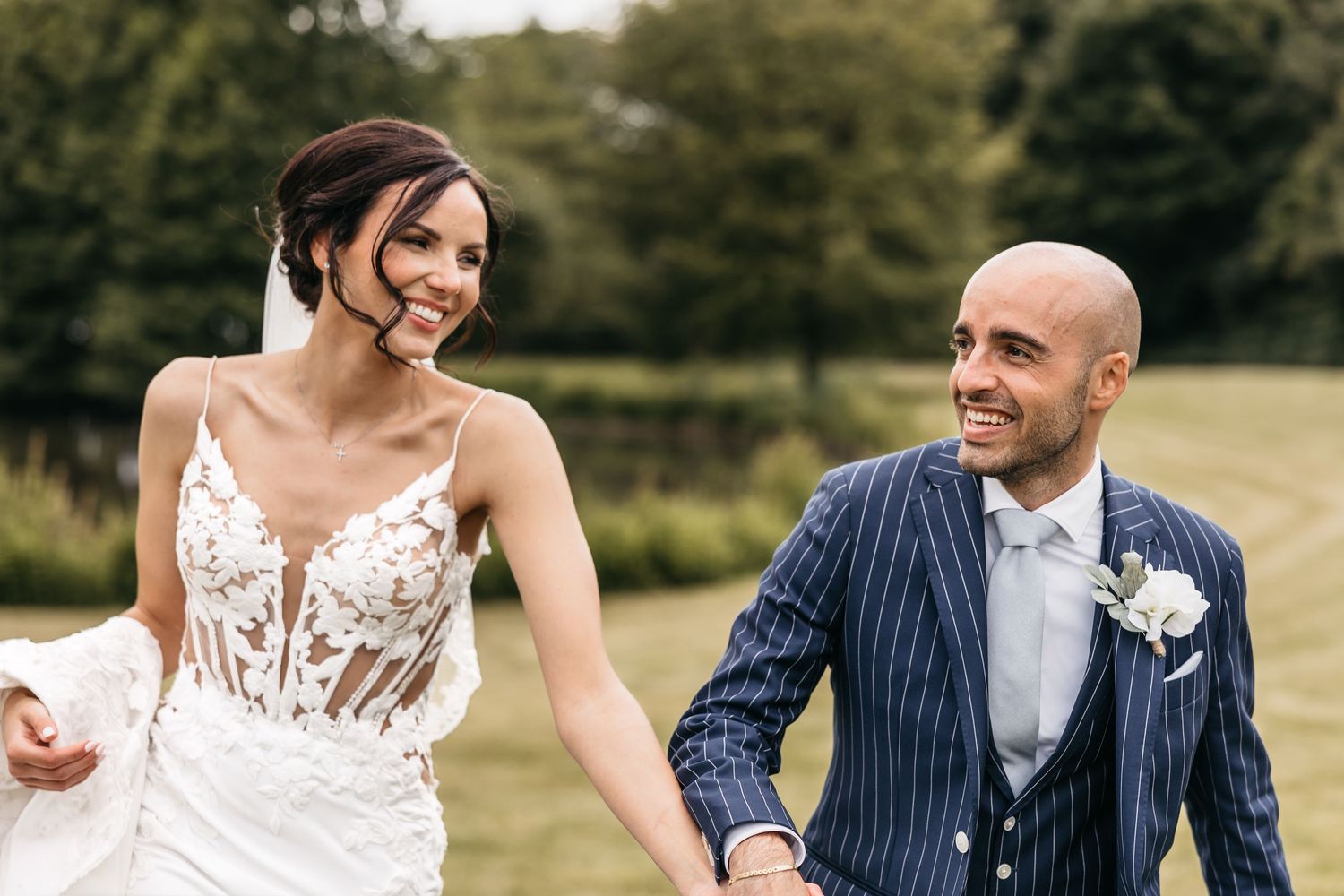 Smiling bride in lace wedding dress and bald groom in navy pinstripe suit walk hand-in-hand outdoors.