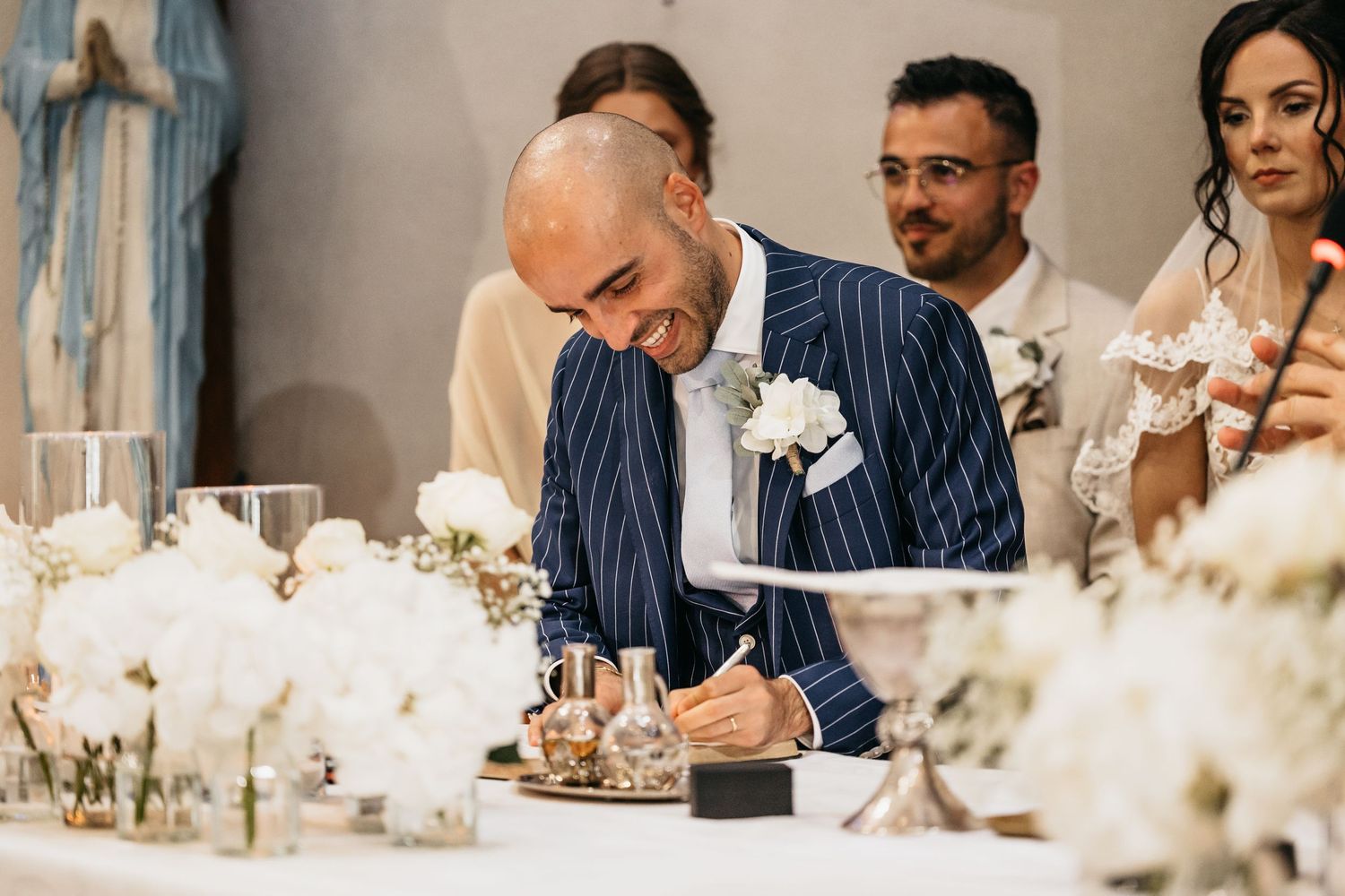 Smiling groom in navy pinstripe suit signing wedding register at altar decorated with white flowers.