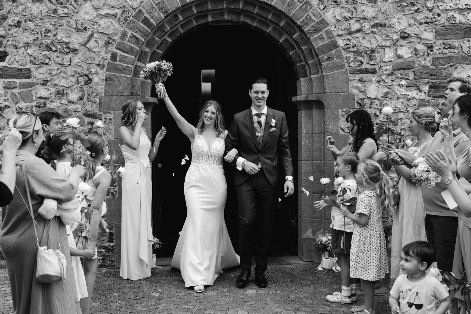 Black and white photo of bride and groom leaving stone church amid celebrating wedding guests.