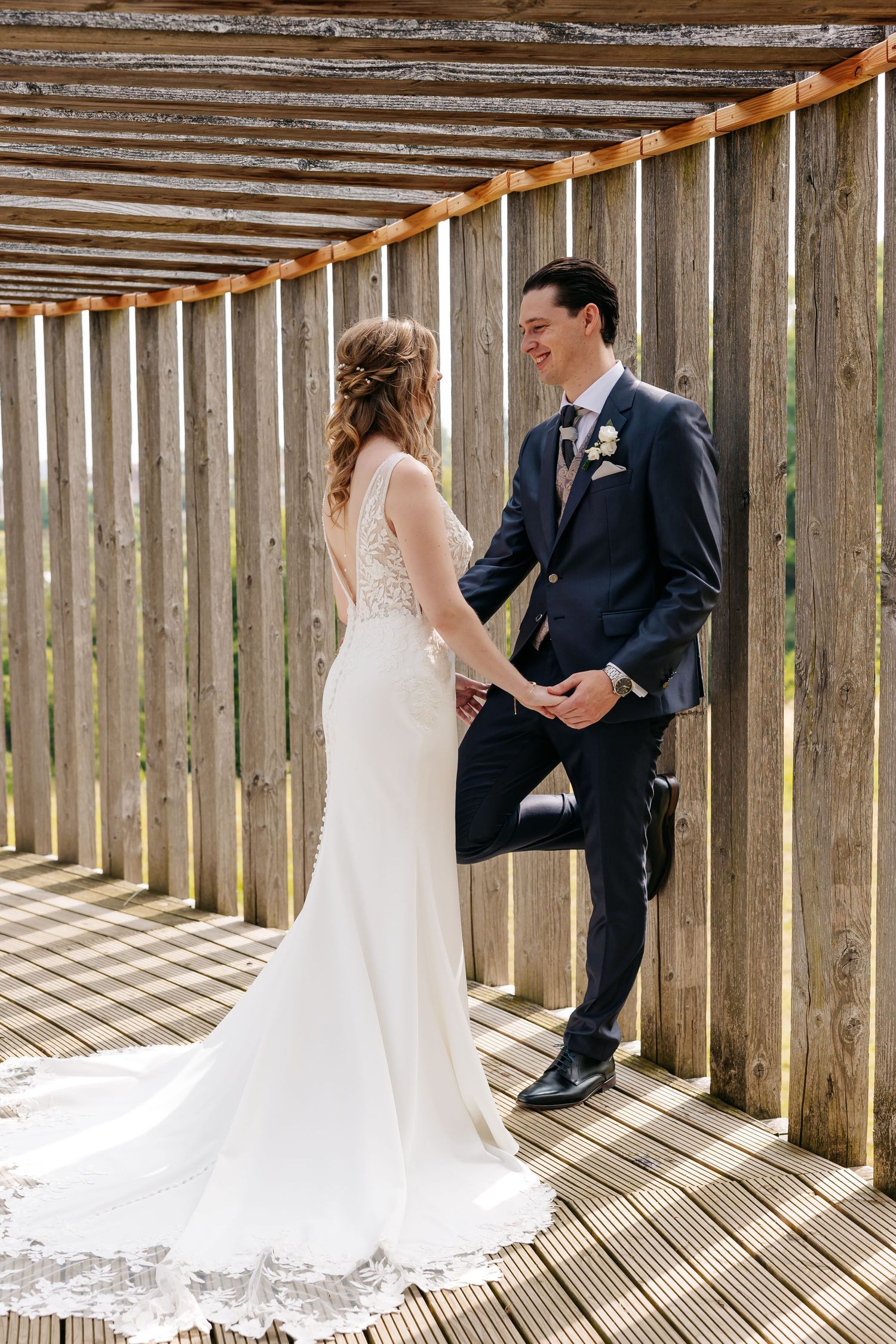 Bride and groom hold hands smiling at each other against rustic wooden fence.