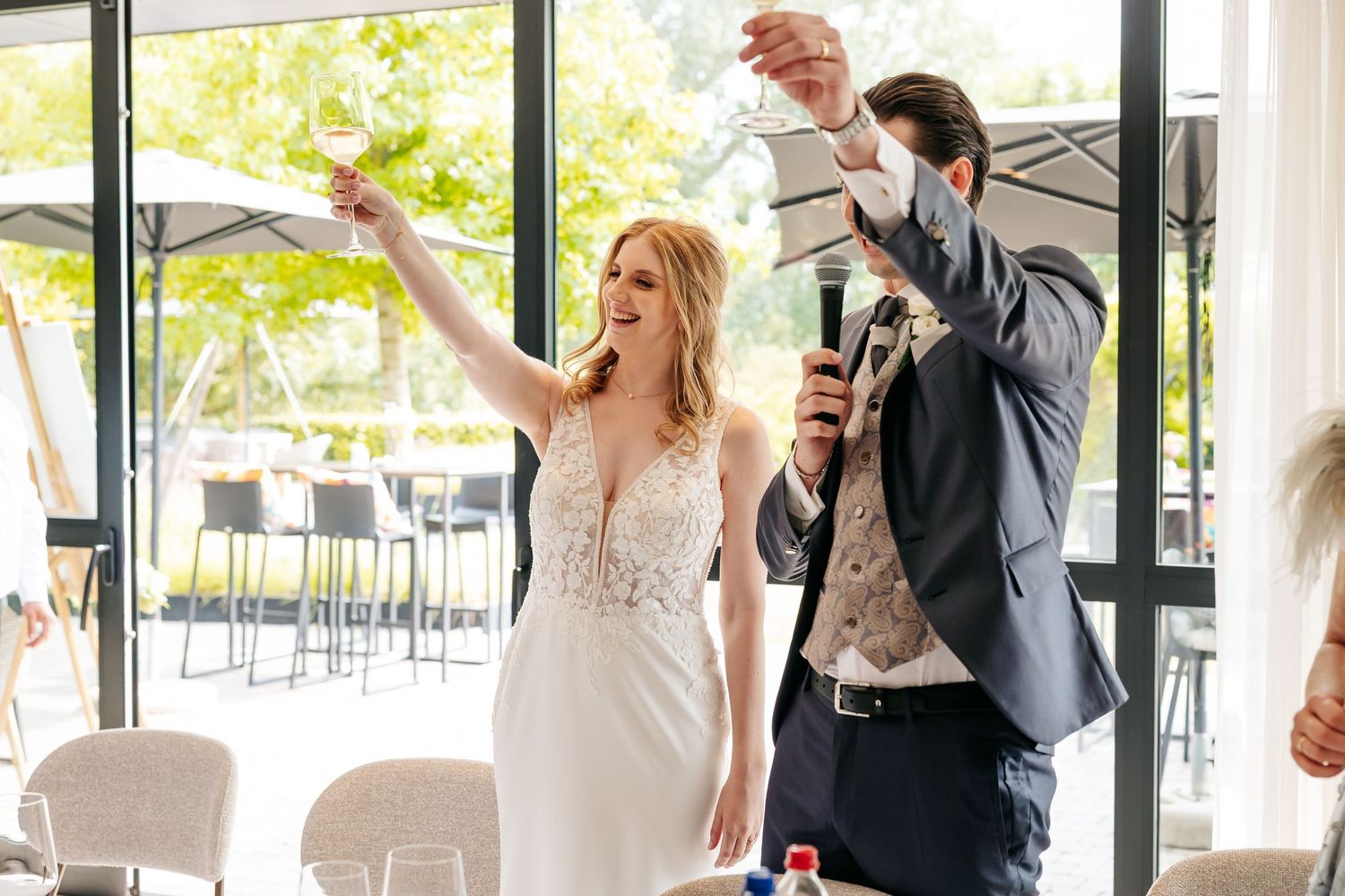 Bride raises glass while groom speaks into microphone during joyful wedding reception toast.