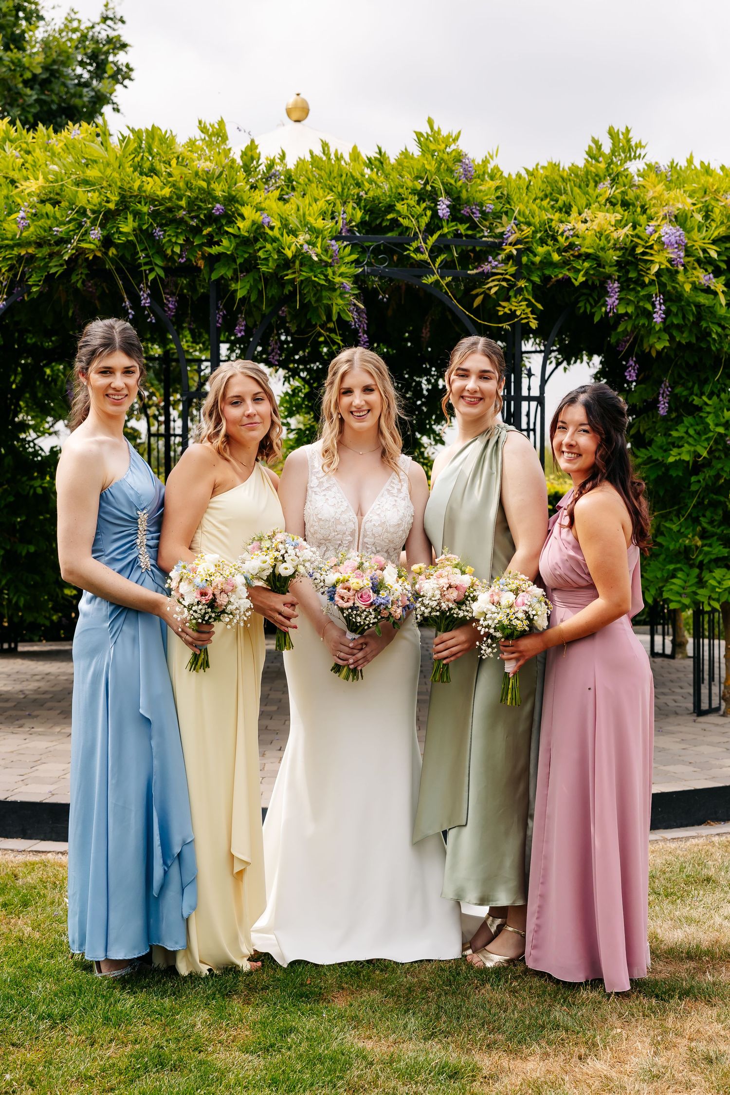 Bride and four bridesmaids in blue, yellow, green, and pink gowns pose with bouquets before a floral arch.