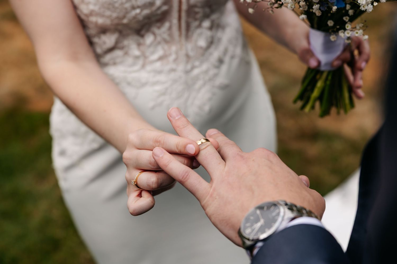 Close-up of groom holding bride's hand with gold wedding band during outdoor wedding ceremony.