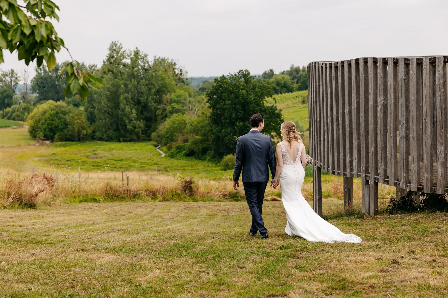 Bride and groom walking hand in hand across a green field beside a rustic wooden fence on their wedding day.