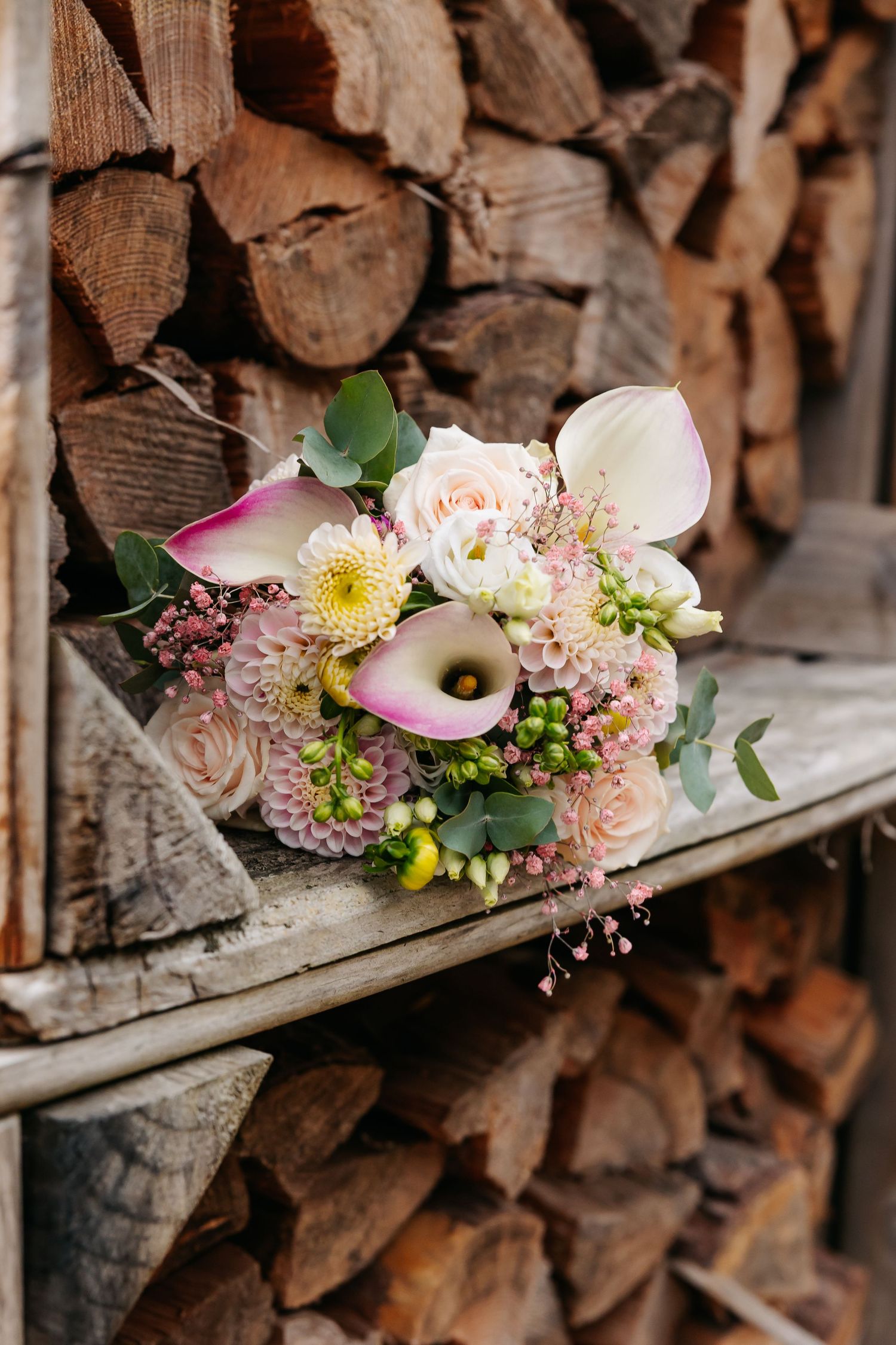 Elegant bridal bouquet with pink roses, calla lilies, and dahlias resting on rustic wooden logs.