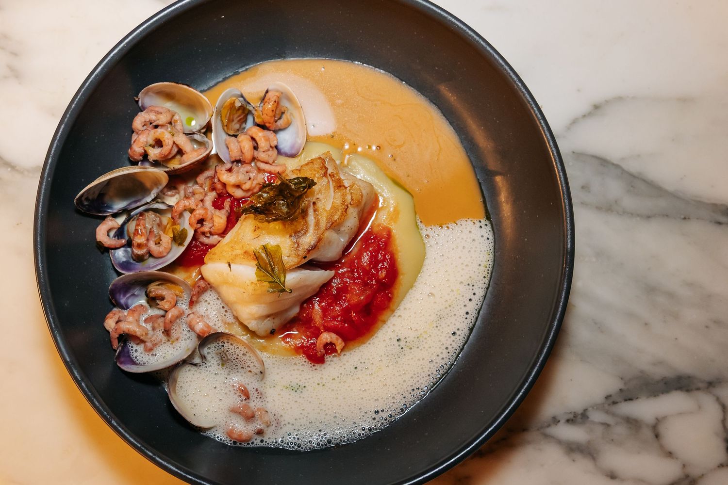 Elegant seafood dish with clams, shrimp, and fish in creamy orange bisque broth in black bowl.