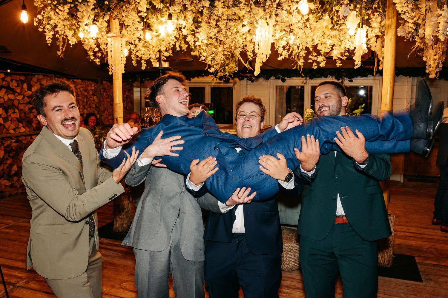 Groomsmen joyfully lifting groom in blue suit during lively wedding reception celebration indoors.