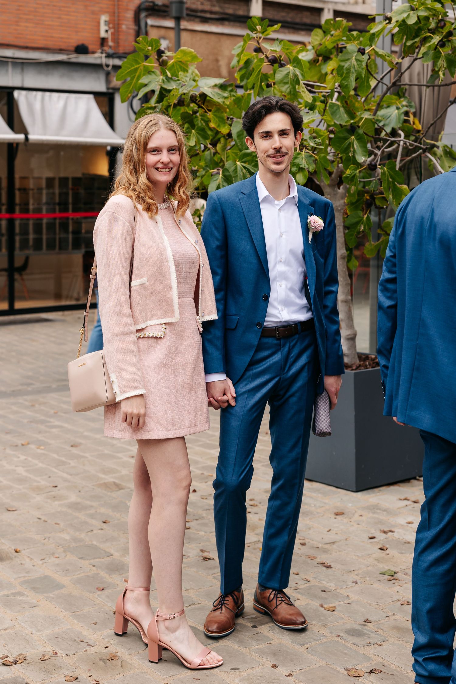 Elegant couple posing outdoors, woman in pink suit dress and man in blue suit with boutonniere at a wedding.