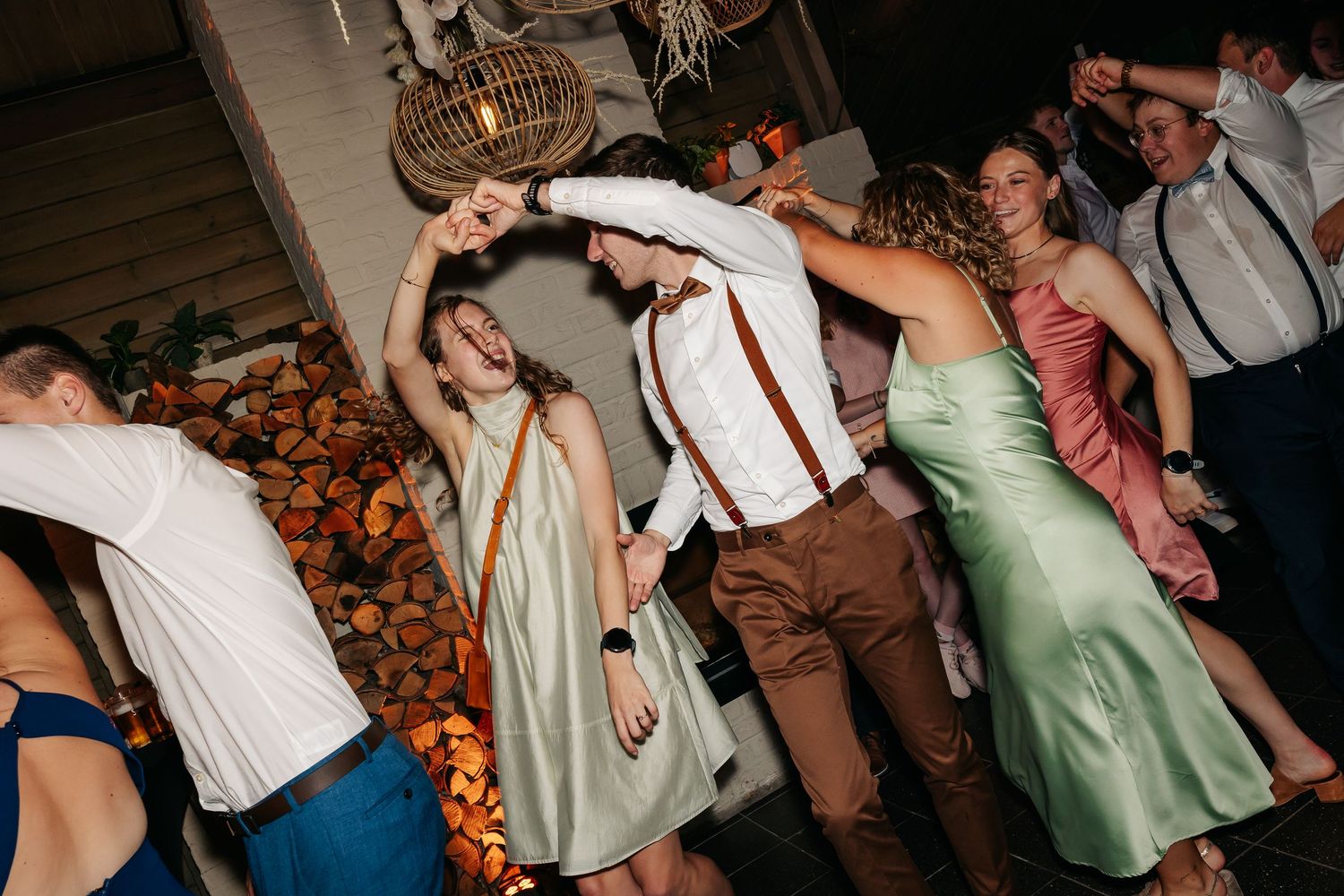 Wedding guests dancing joyfully, groom in suspenders twirling bride in green dress at reception.