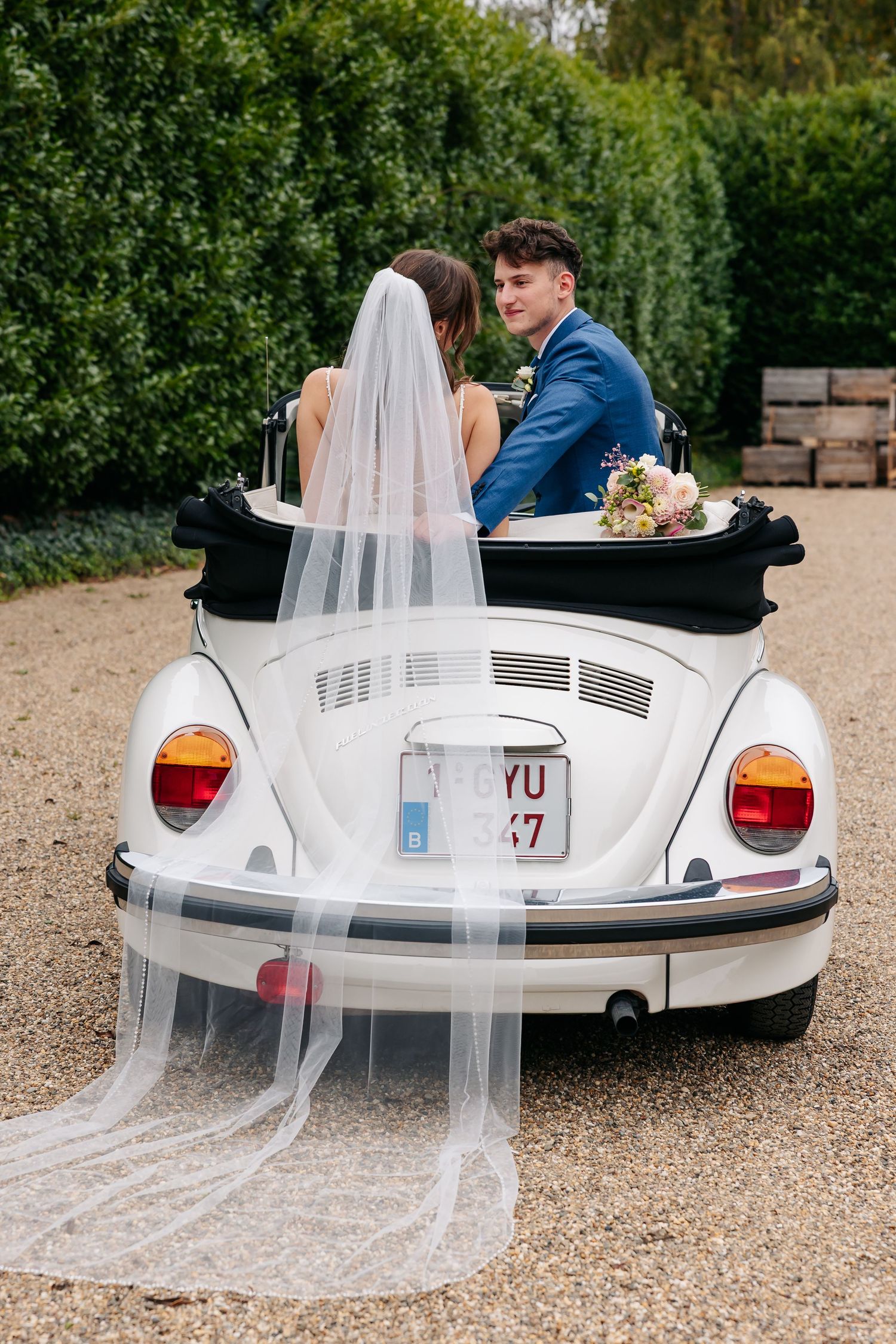 Groom in blue suit turns to smile while seated with bride in white VW Beetle convertible wedding car.