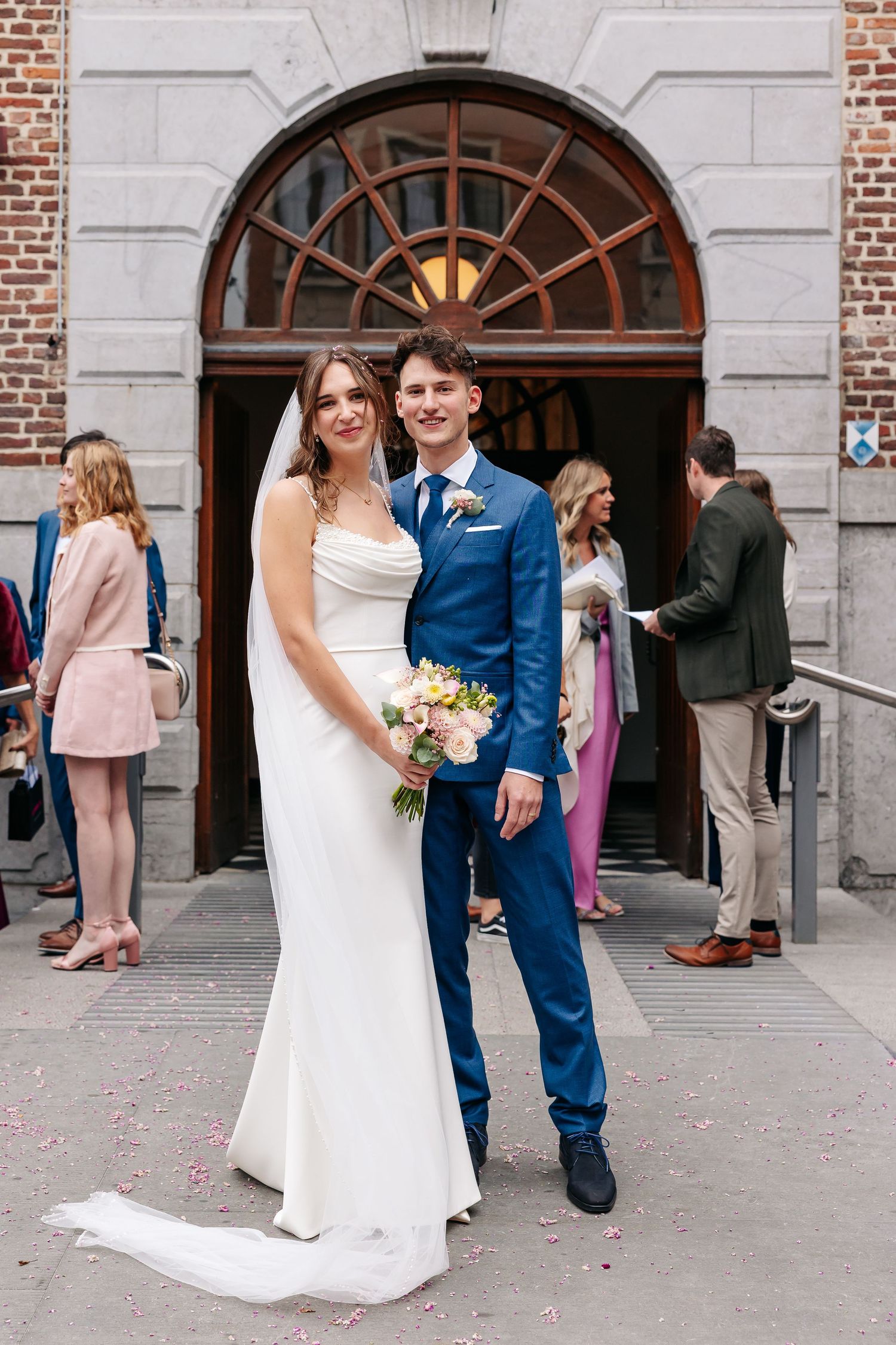 Bride and groom pose together outside church entrance, bride holding pastel bouquet, groom in blue suit.