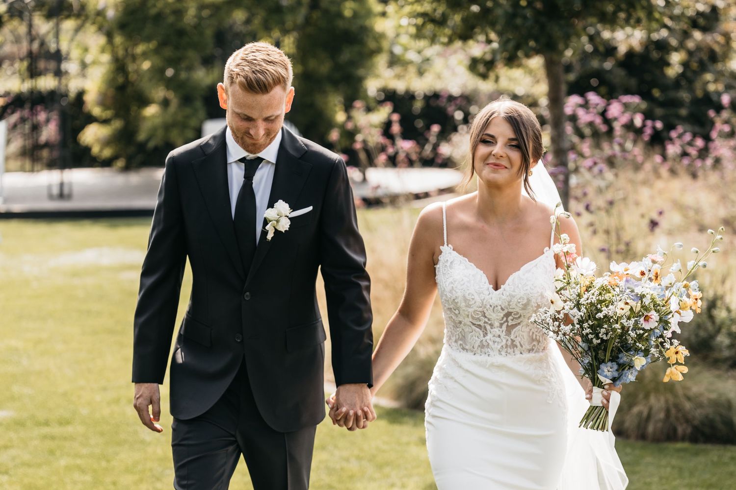Smiling bride in lace wedding gown holds colorful bouquet while walking with groom in black suit.