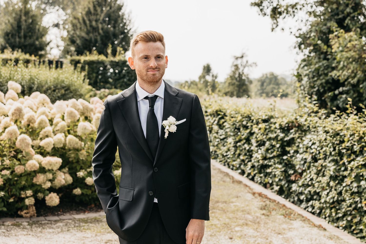 Handsome groom in black tuxedo with white boutonniere standing on a garden path.