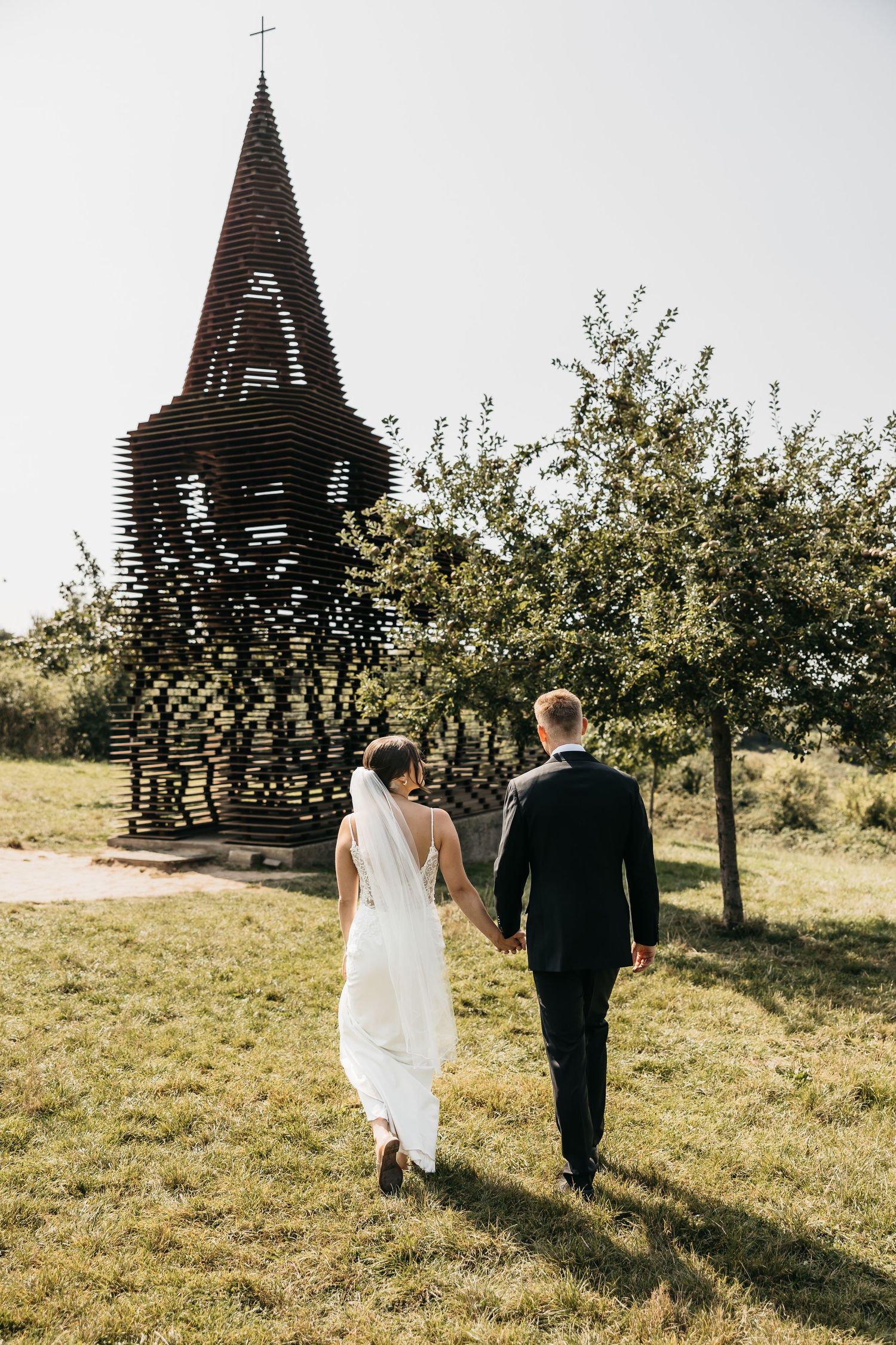 Bride and groom walking hand in hand toward a unique latticed church tower sculpture outdoors.