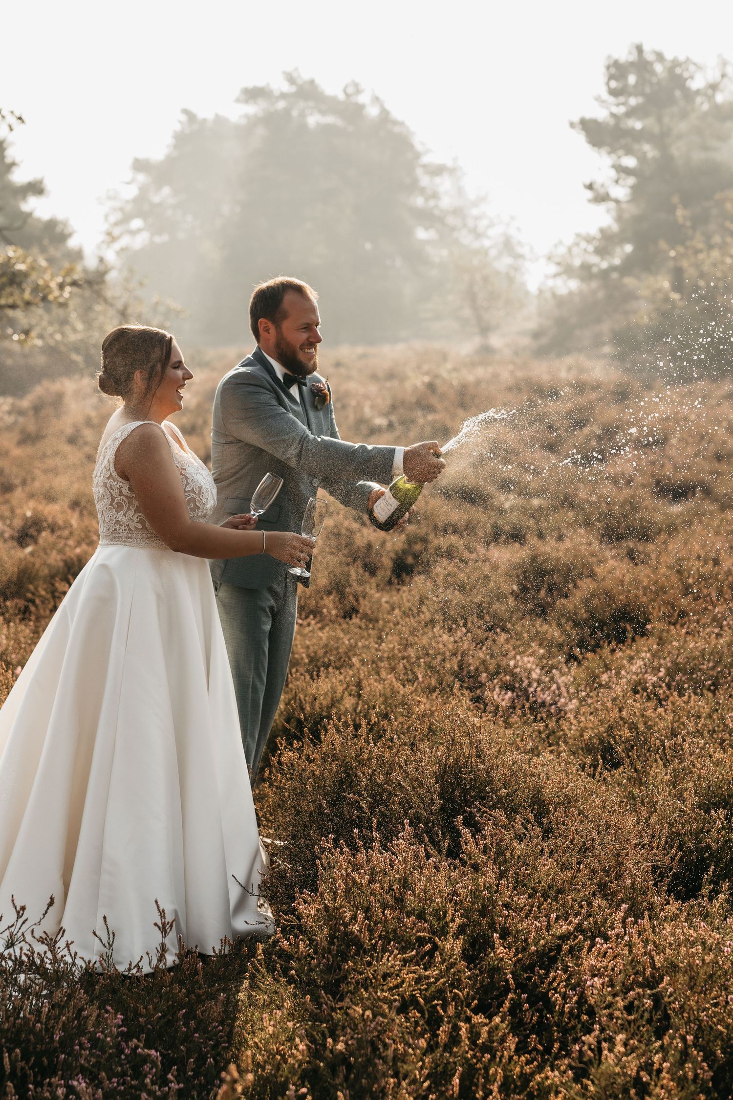 Groom spraying champagne as bride holds a glass in a glowing heather field on wedding day.