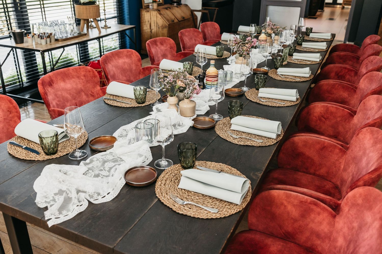 Elegantly set long wedding reception table with red velvet chairs, woven placemats, and floral centerpieces.