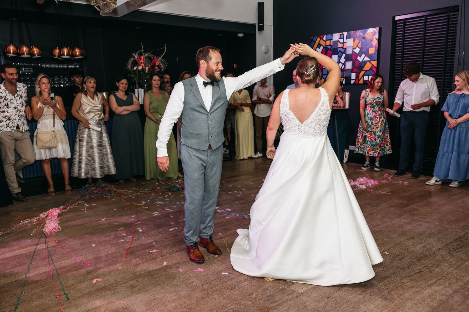Groom twirls his bride in a stunning white ball gown during their first dance at a colorful wedding reception.