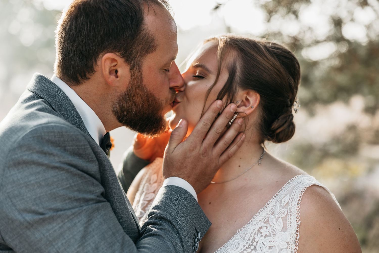 Bride and groom share a passionate kiss with the groom's hand gently cupping her face outdoors.