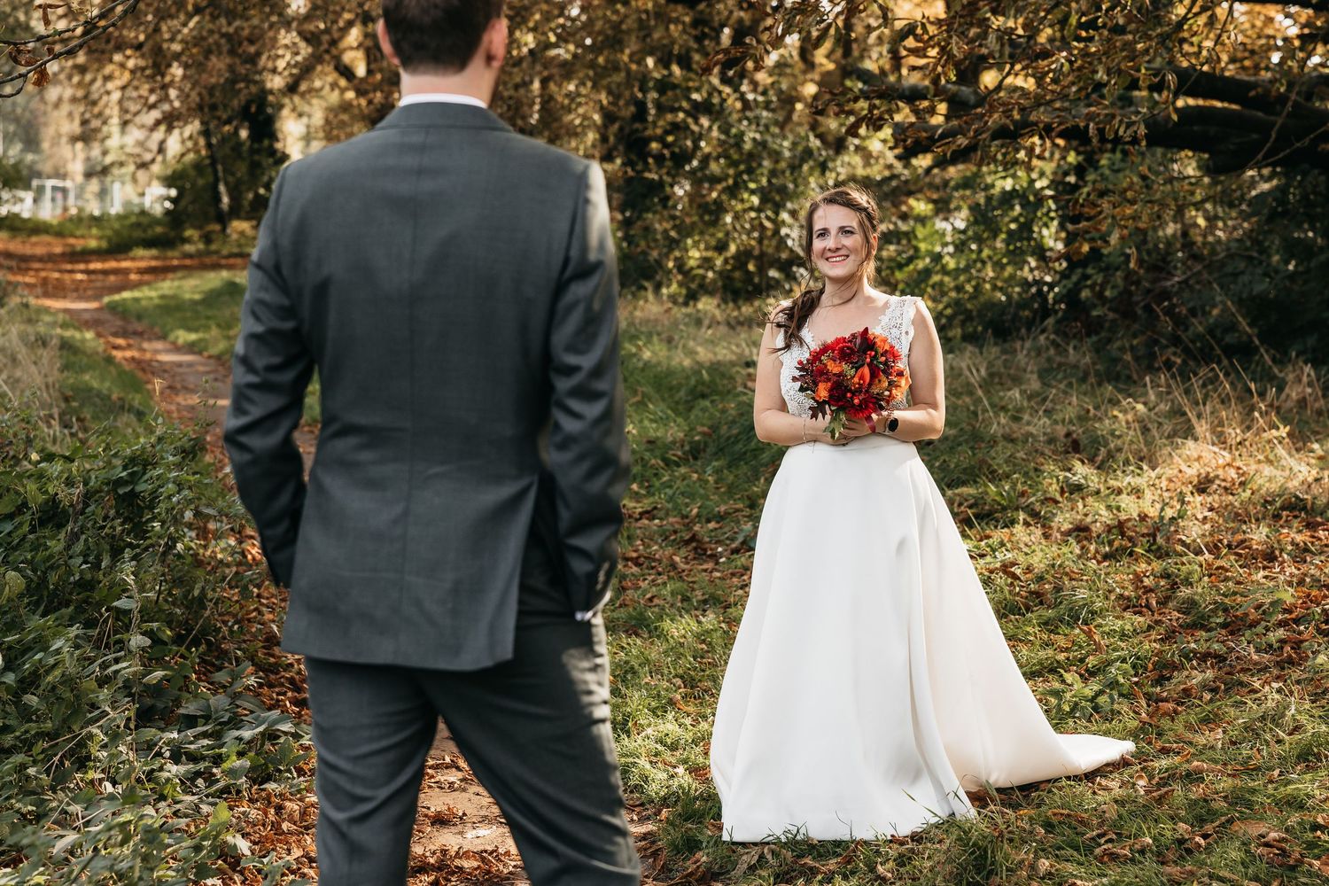 Smiling bride in white gown holding red bouquet facing groom in gray suit in autumn woods.
