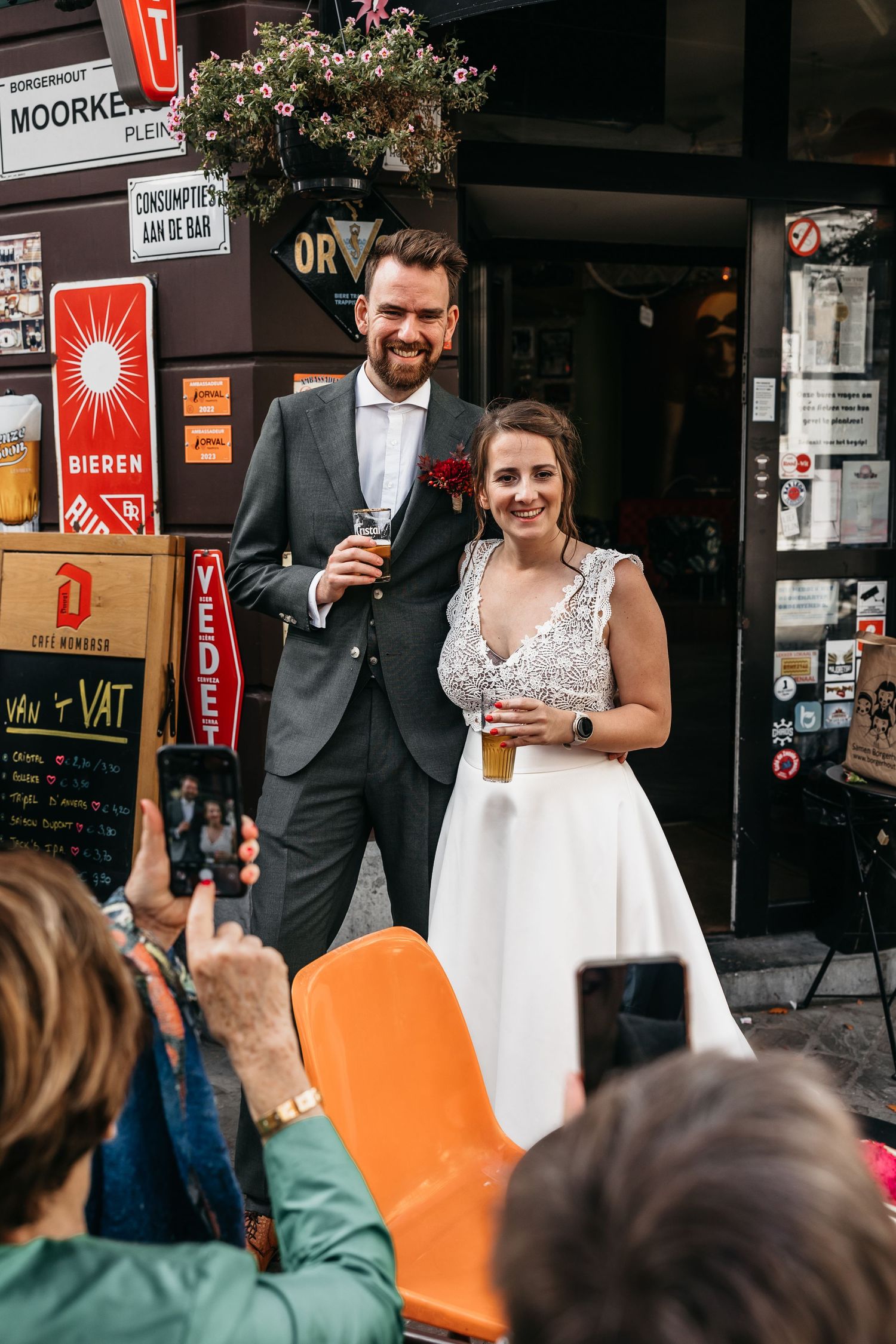Smiling bride and groom holding drinks while guests photograph them outside a Belgian café.