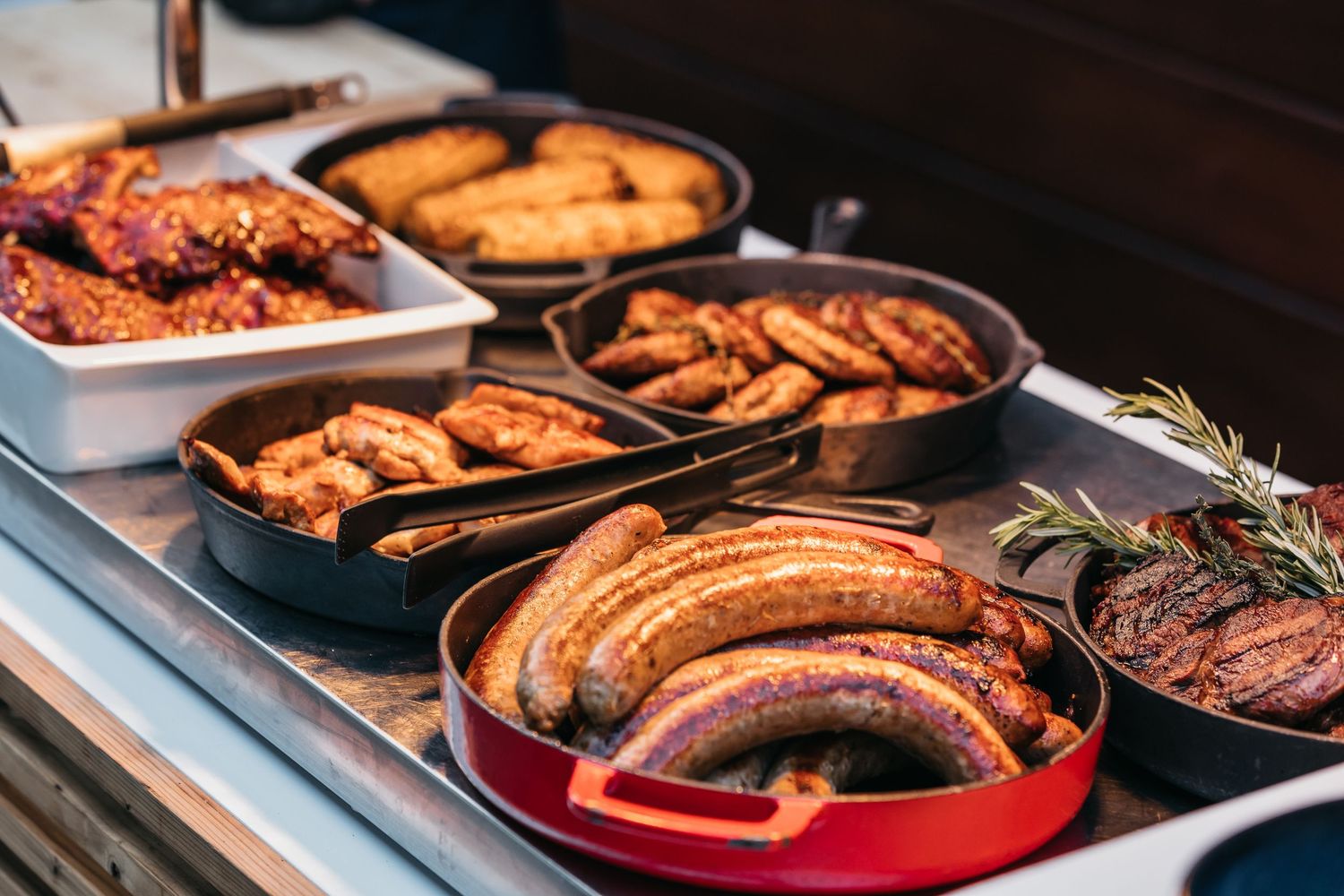 Assorted grilled sausages and meats displayed in cast iron pans at an outdoor buffet station.