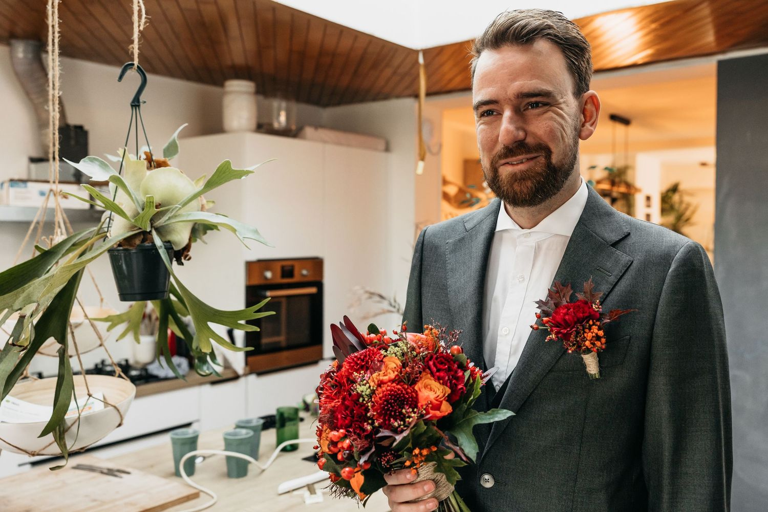 Handsome groom in grey suit holding a stunning red and orange floral bouquet with a boutonniere.