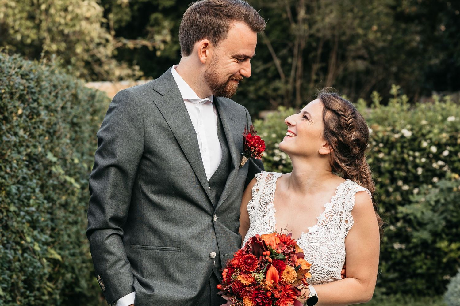 Happy newlyweds sharing a loving gaze, bride holding red bouquet in front of green hedges.
