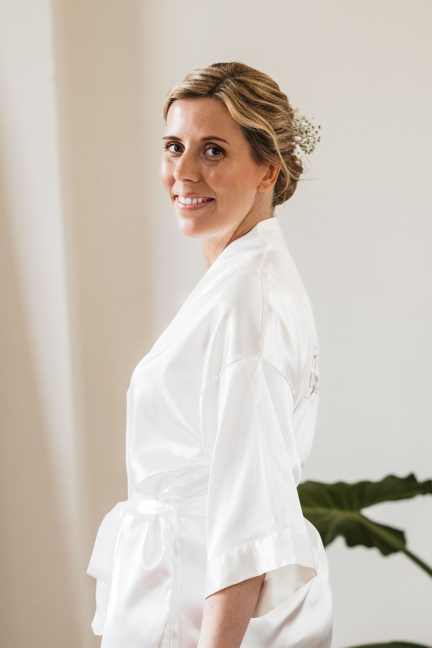 Elegant bride in a white satin robe smiling, with delicate floral hair accessories in an updo.