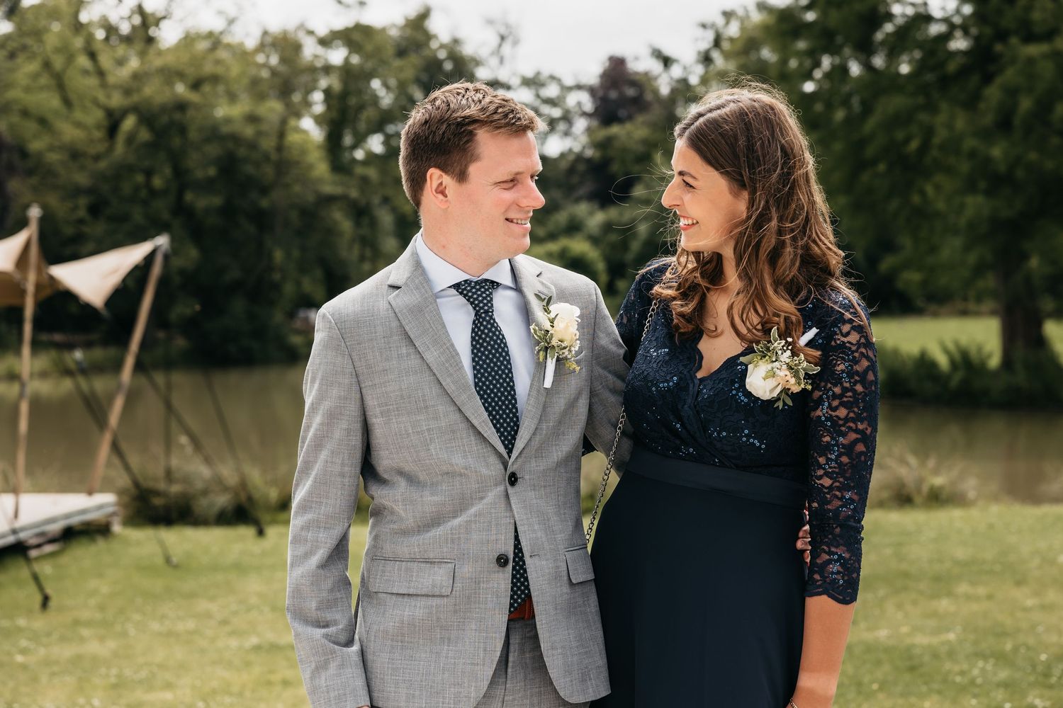 Man in grey suit and woman in navy lace dress smile at each other at an outdoor garden wedding.