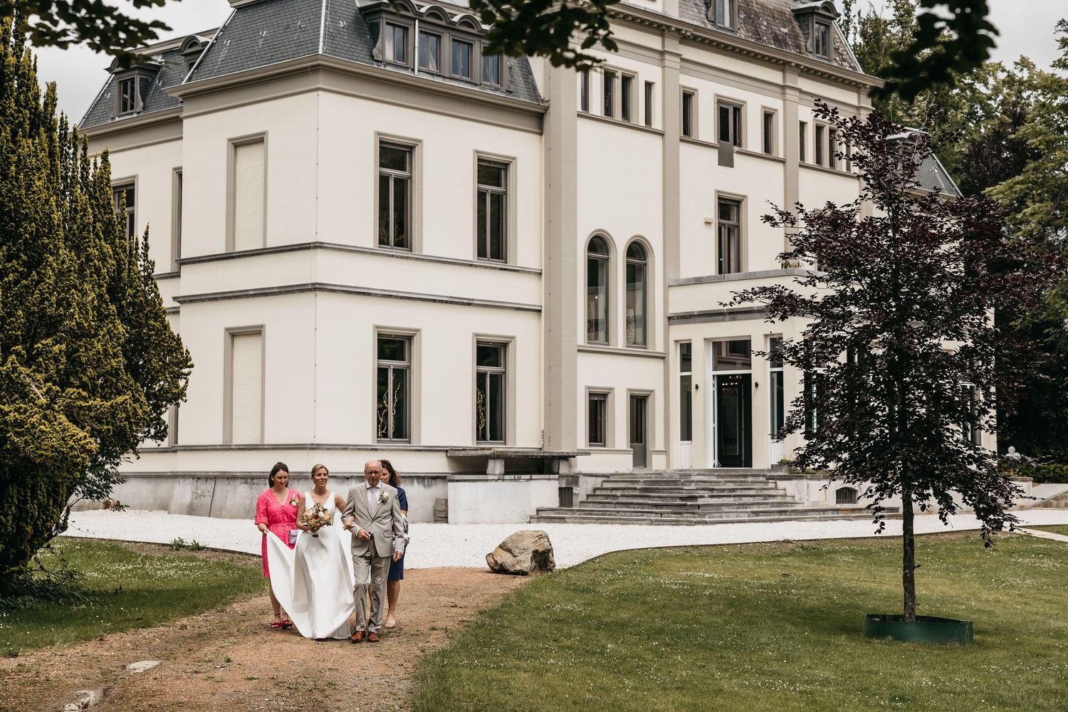 Bride in white gown walks with two guests in pink and grey outfits outside a white chateau wedding venue.